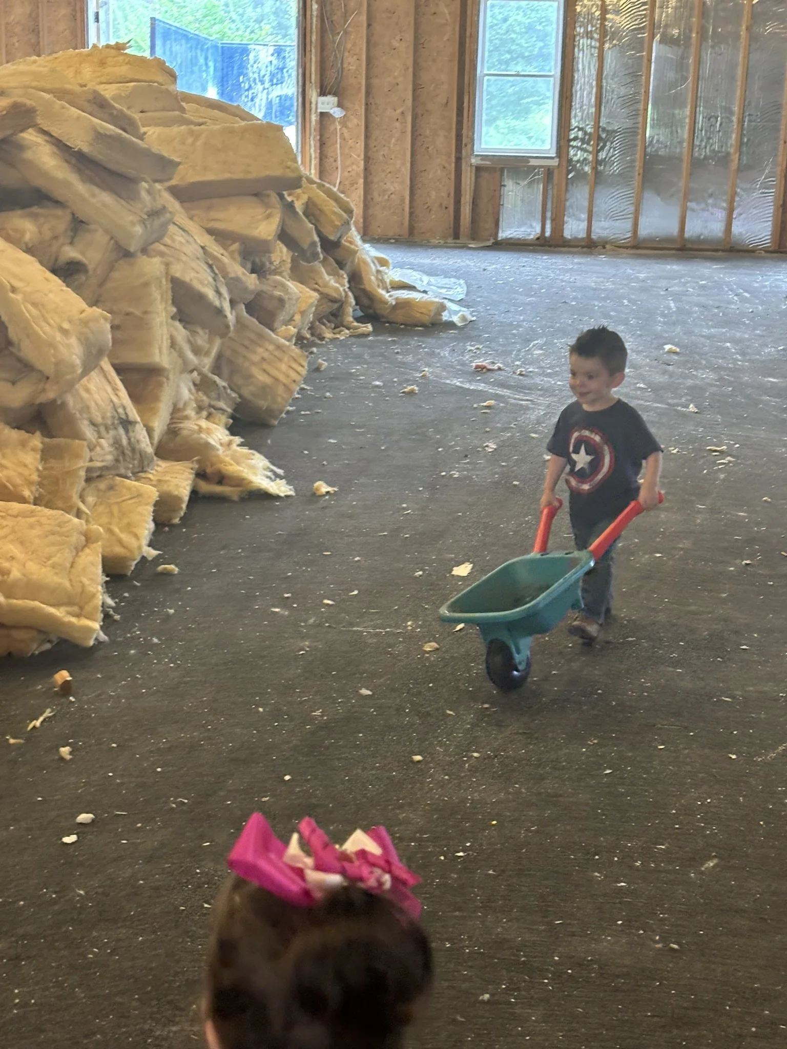 A young boy wearing a Captain America T-shirt pulls a small green wheelbarrow in an unfinished room with piles of yellow insulation on the floor and exposed wooden framing on the walls and ceiling.