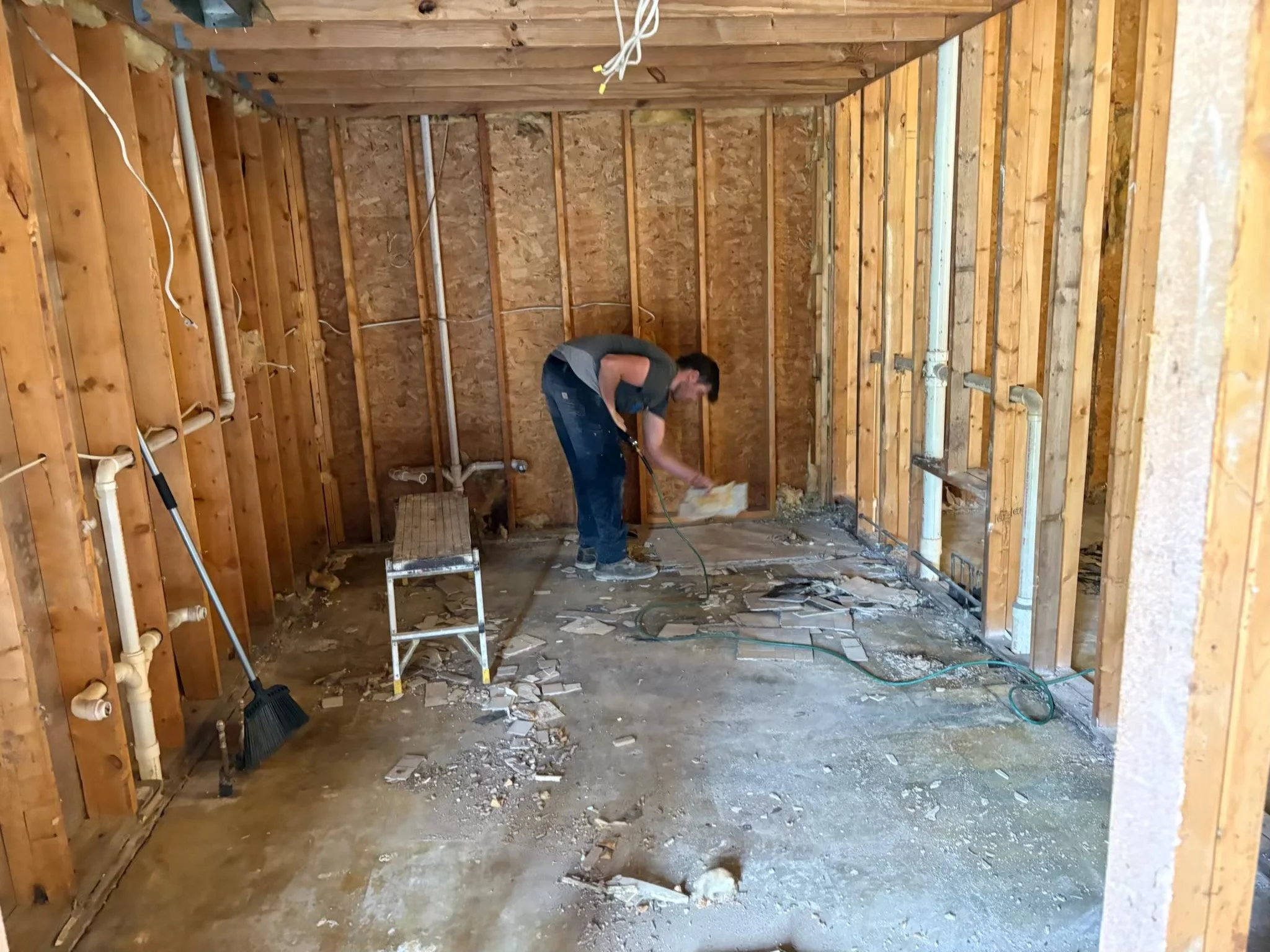 Man working on a construction or renovation project inside a wooden-framed room. The room has exposed pipes and framing with debris on the floor.