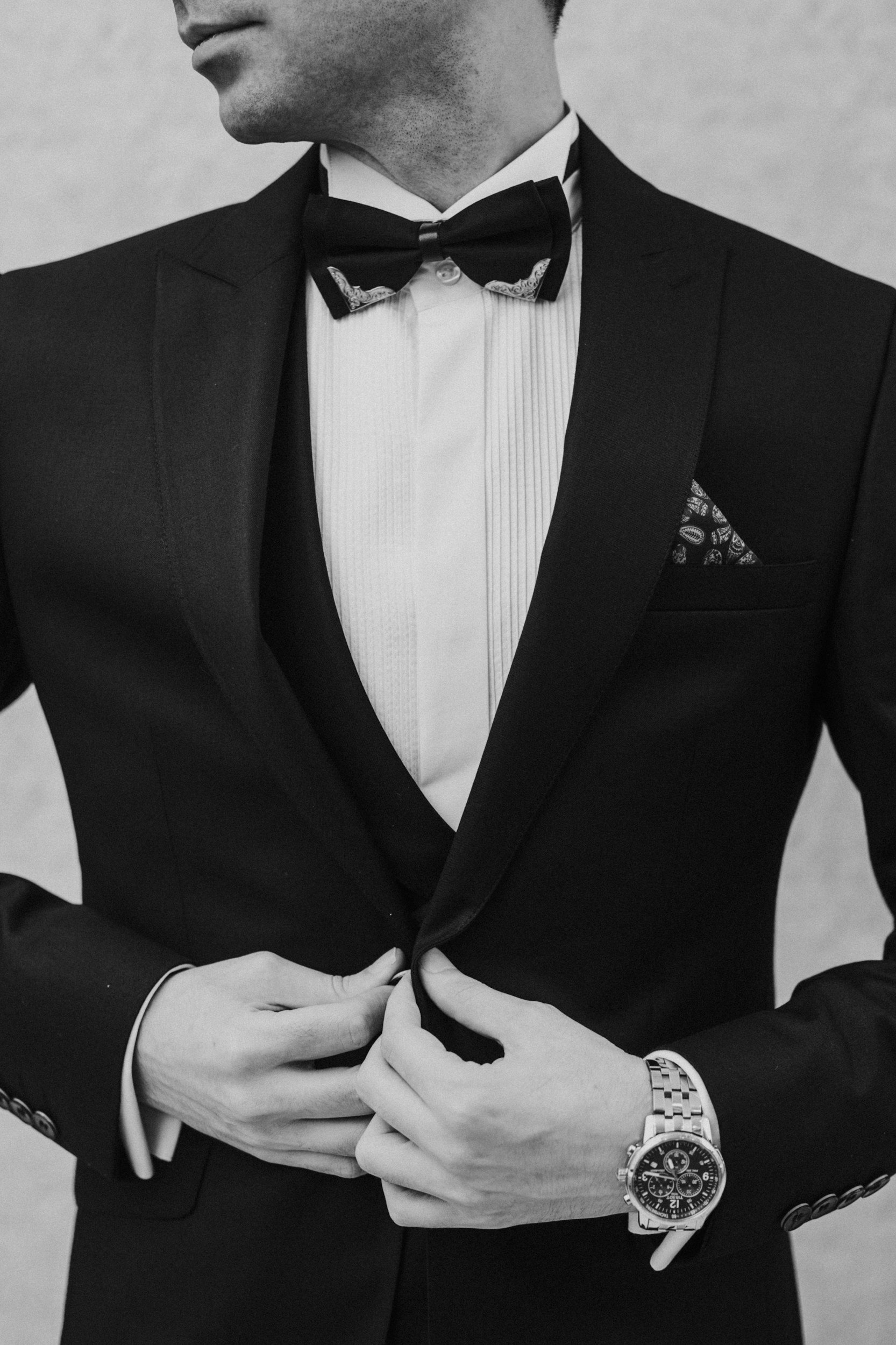 Close-up of a man in a tuxedo adjusting his cuffs, wearing a watch and a bowtie, with a patterned pocket square visible.