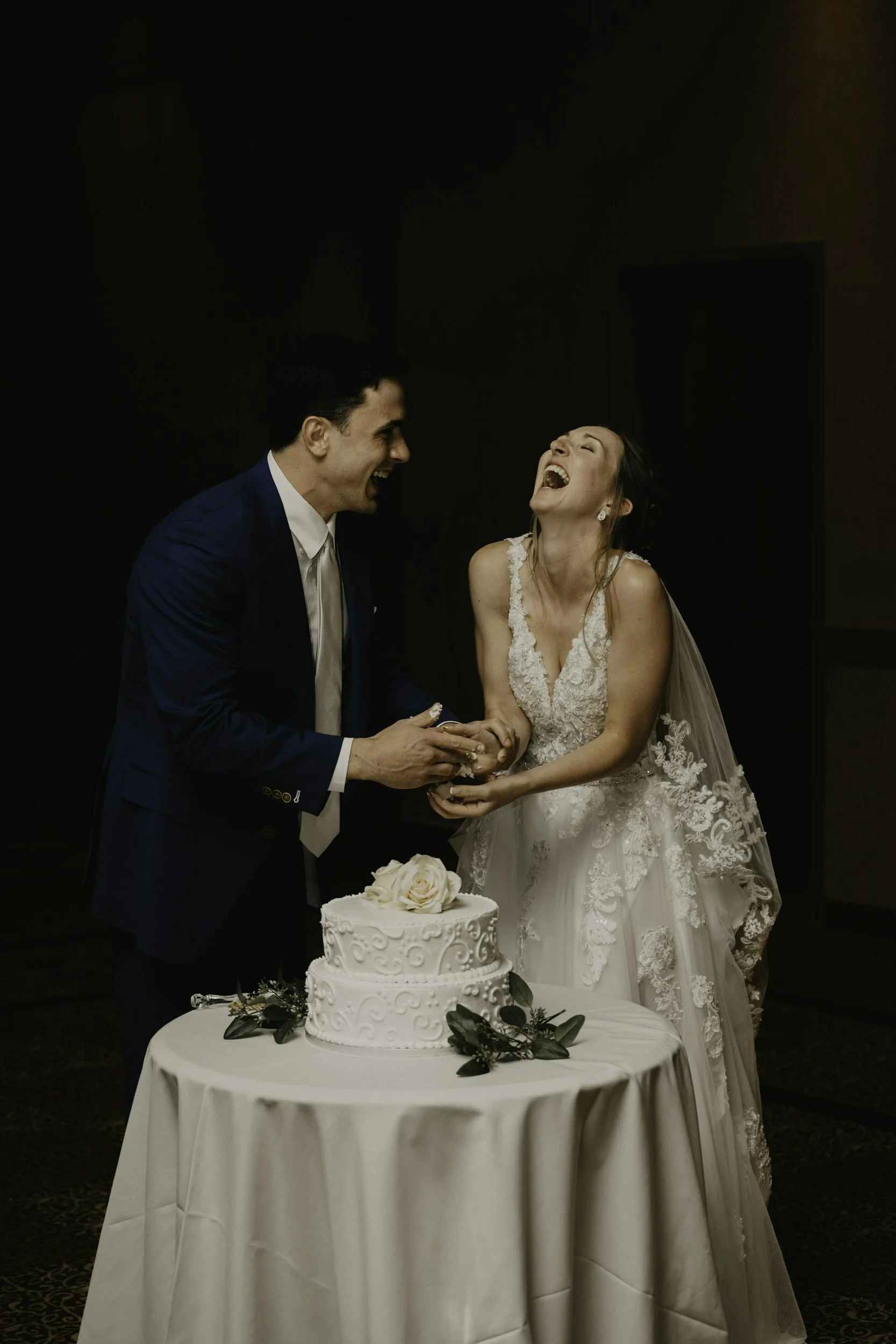 A bride and groom laughing during their wedding celebration, standing behind a wedding cake decorated with roses and greenery.