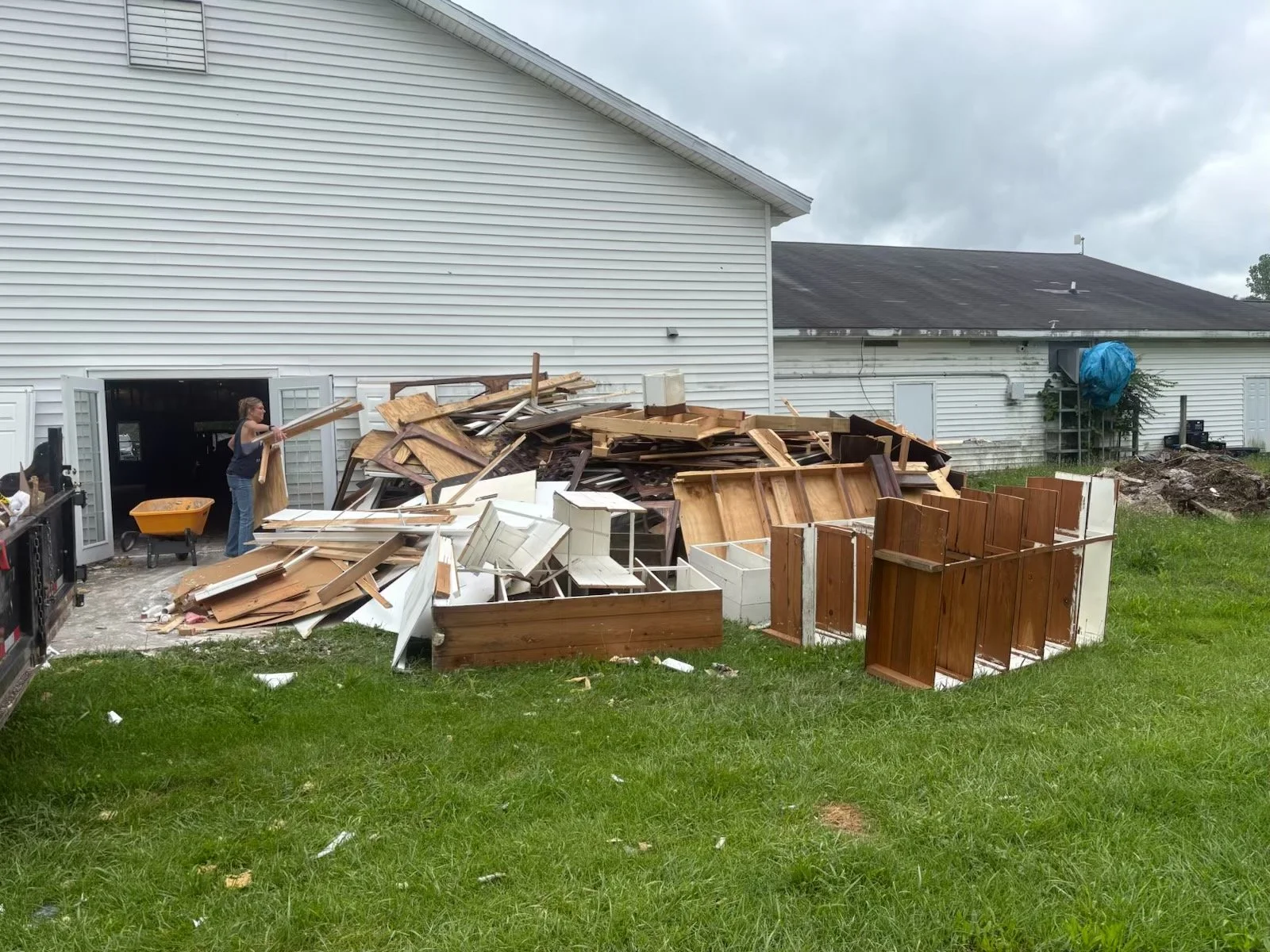 A woman stacking wood and moving debris outside a house with a large pile of broken furniture, wood, and debris in the yard on a cloudy day.