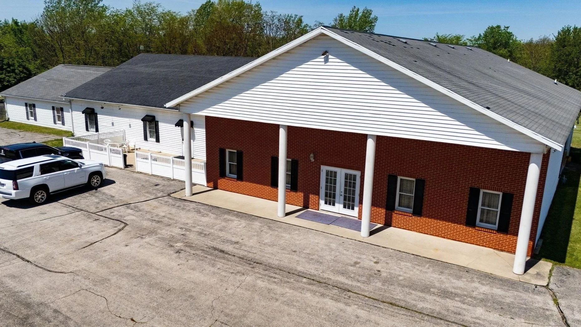 An aerial view of a building with white and red brick exterior, black shutters, white columns, and a gray shingled roof, adjacent to a parking lot with several parked vehicles.