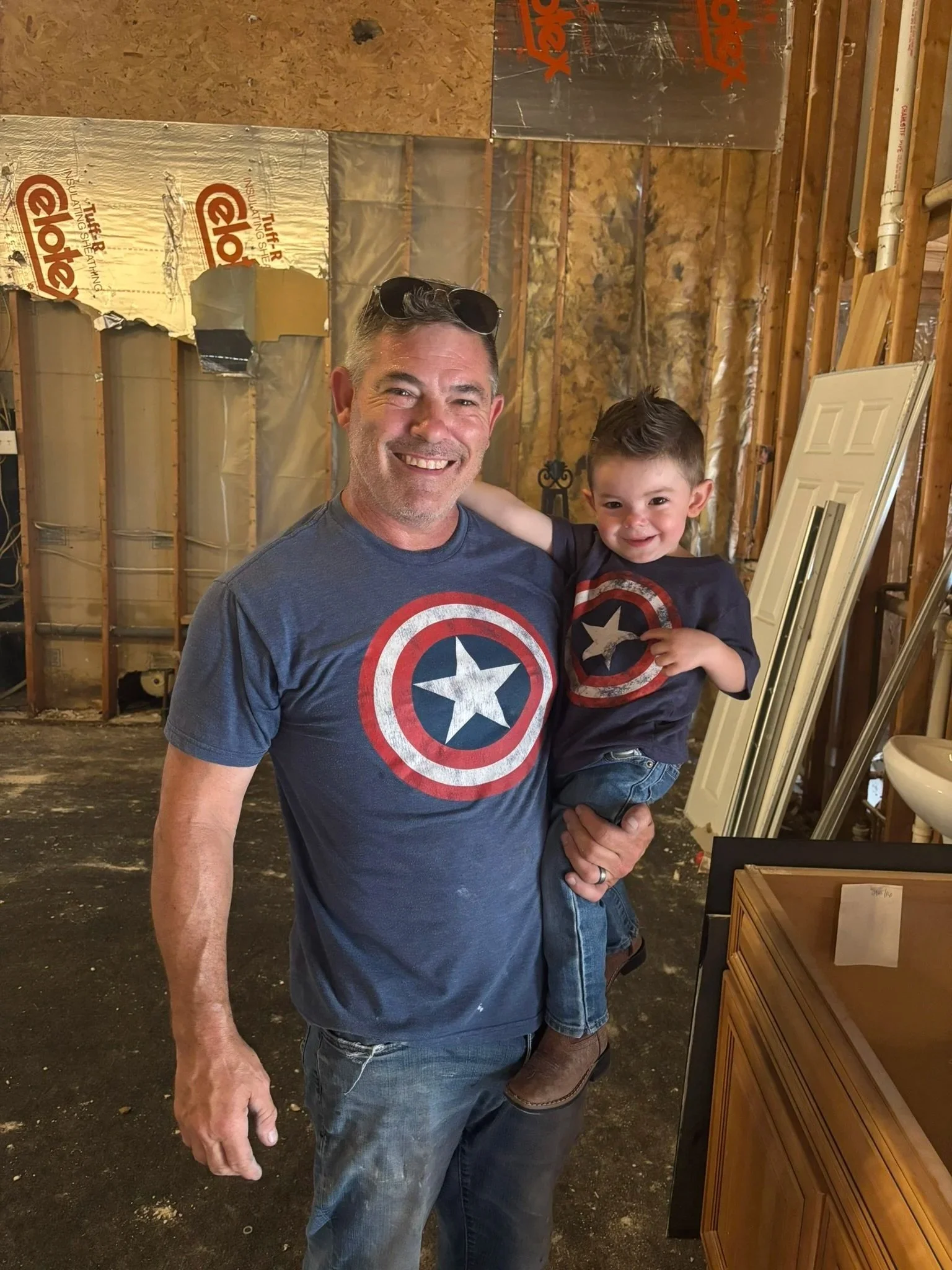 A smiling adult man with sunglasses on his head and a young boy with a Captain America t-shirt, in a room with unfinished walls and some furniture.