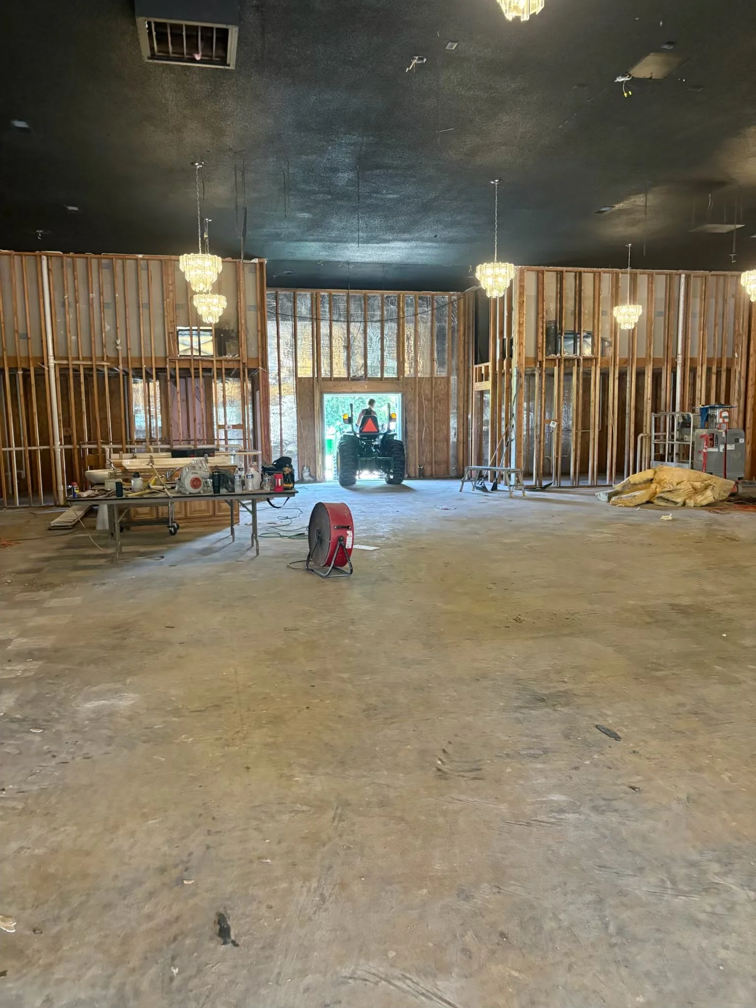Interior of a building under construction with exposed wooden framing, hanging chandeliers, construction tools, and a small tractor near the entrance.