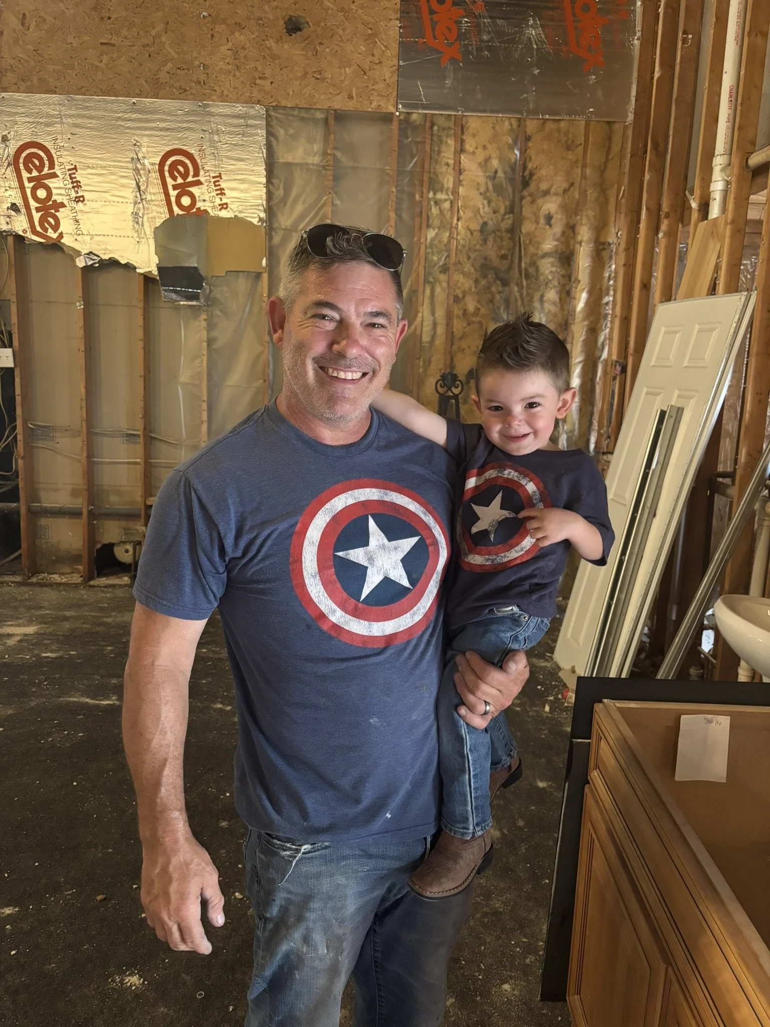 A man holding a young boy, both wearing Captain America T-shirts, smiling in a partially finished room with exposed insulation and wooden framing.