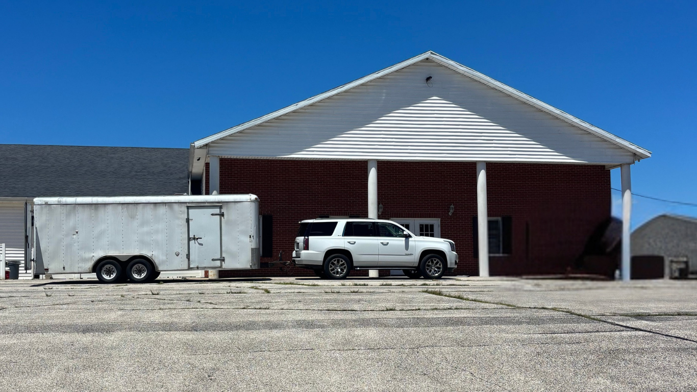 A white SUV parked on a concrete lot in front of a building with white siding and red brick walls, with a white enclosed trailer attached to the SUV, under a blue sky. The truck indicates that the building is under construction.