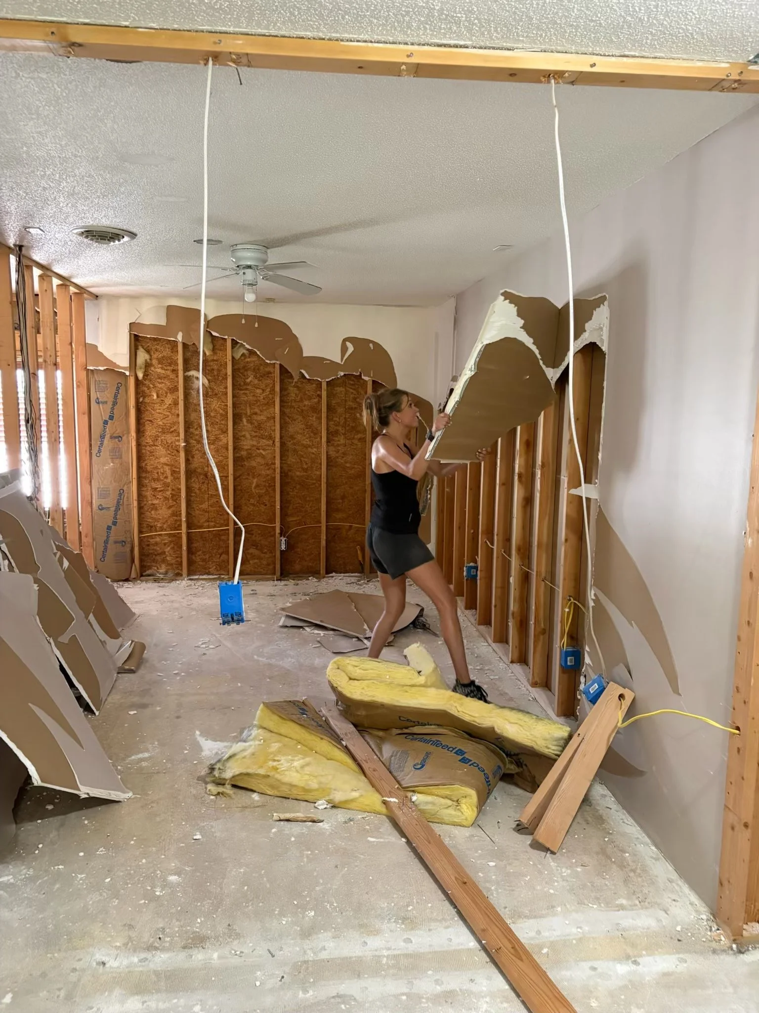 A woman removing drywall from a partially demolished room with exposed wall studs, insulation, and construction debris on the floor.