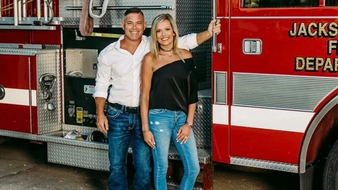 A smiling man and woman standing in front of a red fire truck, with the man wearing a white shirt and jeans and the woman wearing a black off-shoulder top and jeans.