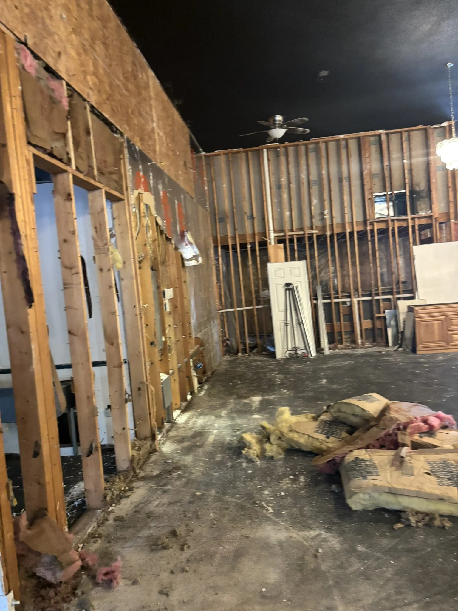 Interior of a room under renovation with exposed wooden wall framing, insulation on the floor, and a ceiling fan.
