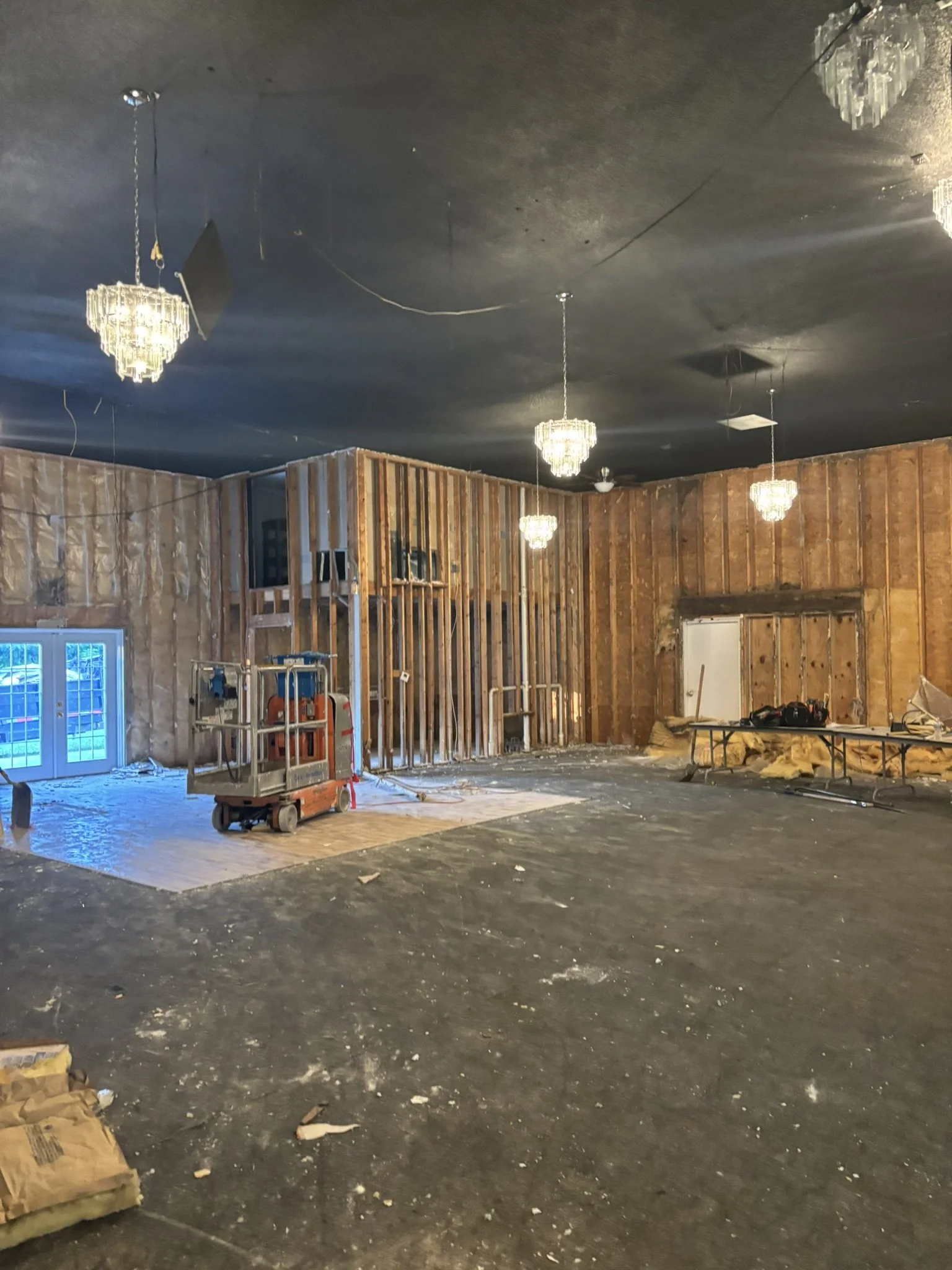 Interior of a room under construction or renovation with exposed wooden wall framing, chandelier ceiling lights, construction equipment, and a partially finished ceiling.