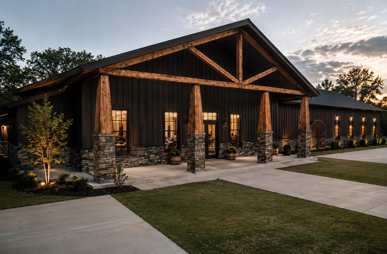 Exterior of a large, modern barn with black siding, stone foundation, and wooden beams under a sunset sky. The barn has multiple windows and a rope-lit pathway leading to the entrance, with landscaped yard and trees nearby.