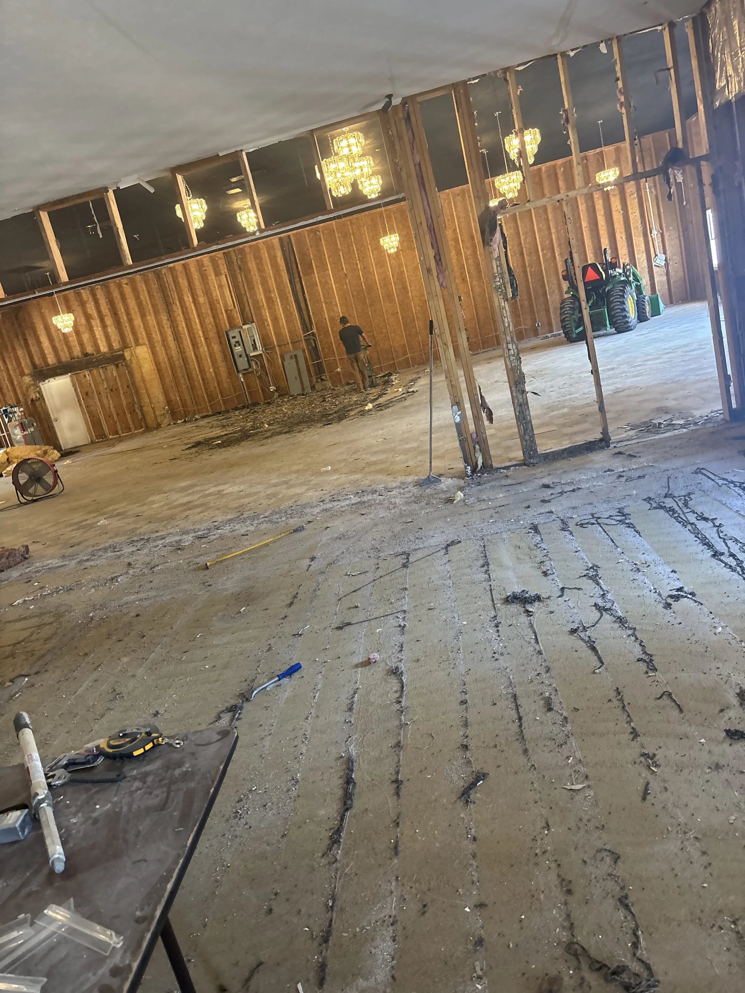 Interior of a building under construction, with exposed wooden framing, hanging chandeliers, a small green tractor, construction tools, and two workers.