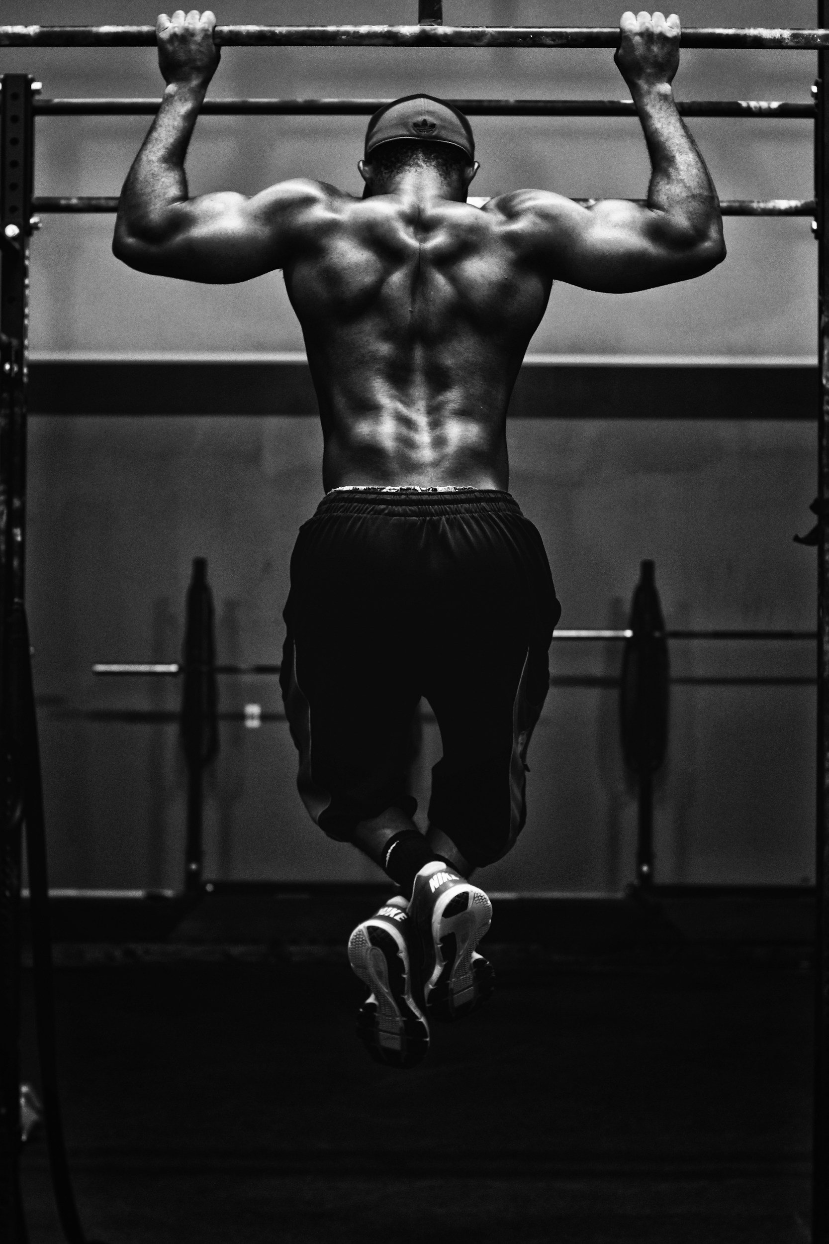 Black and white photograph of a shirtless man doing pull ups in a gym, wearing a backward cap and athletic shoes.