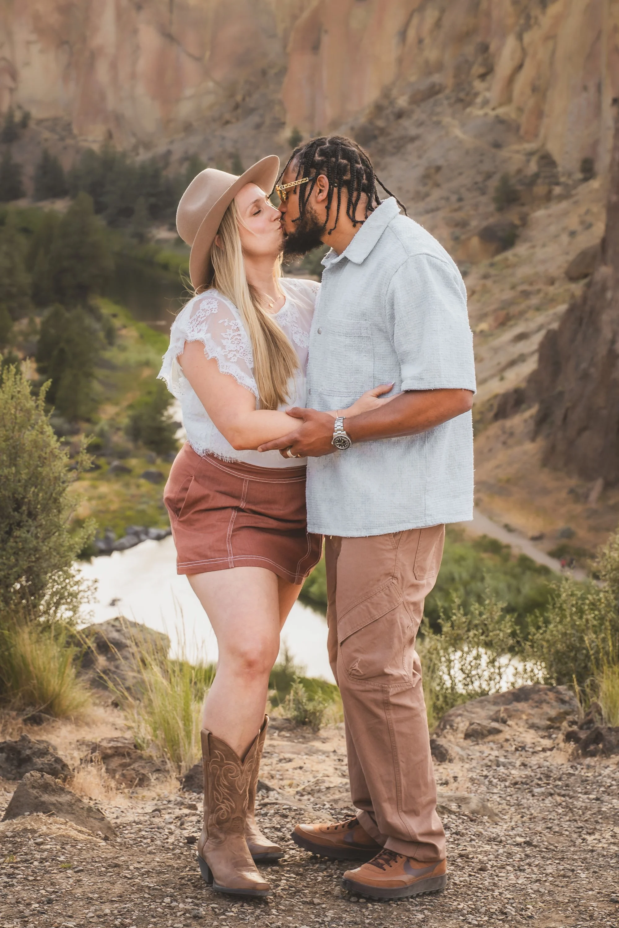 A couple is sharing a kiss outdoors with a canyon and river in the background. The woman is wearing a wide-brimmed hat, lace top, and cowboy boots; the man is in a light-colored shirt, cargo pants, and hiking boots.