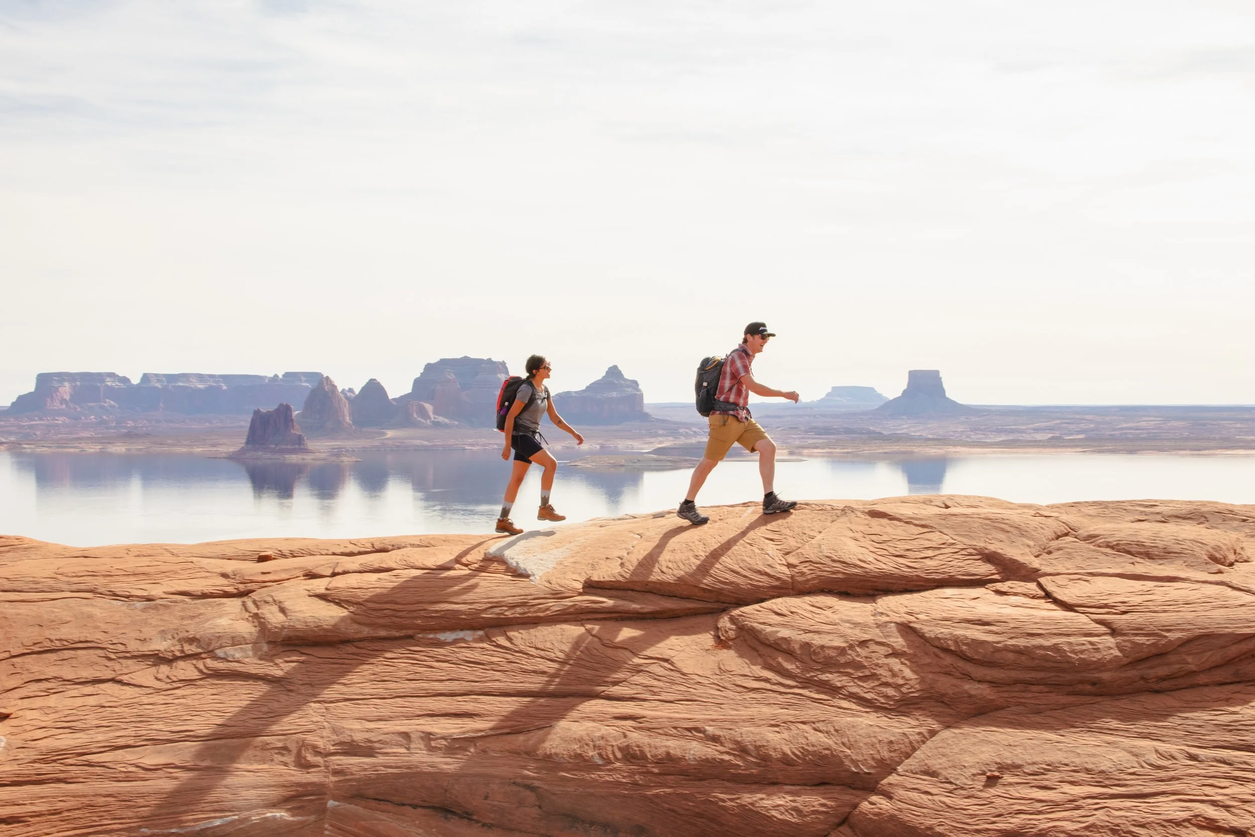 Two hikers, a man and a woman, trekking along a rock formation near a body of water with desert mesas and rock formations in the background under a cloudy sky.