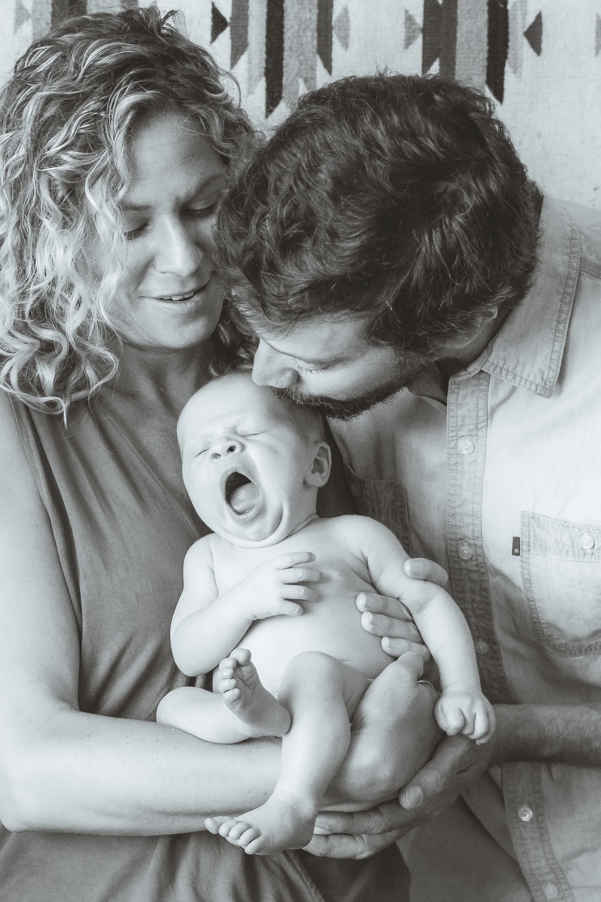 A family holding a baby and sharing a joyful moment, with a woman, a man, and a baby who is yawning.