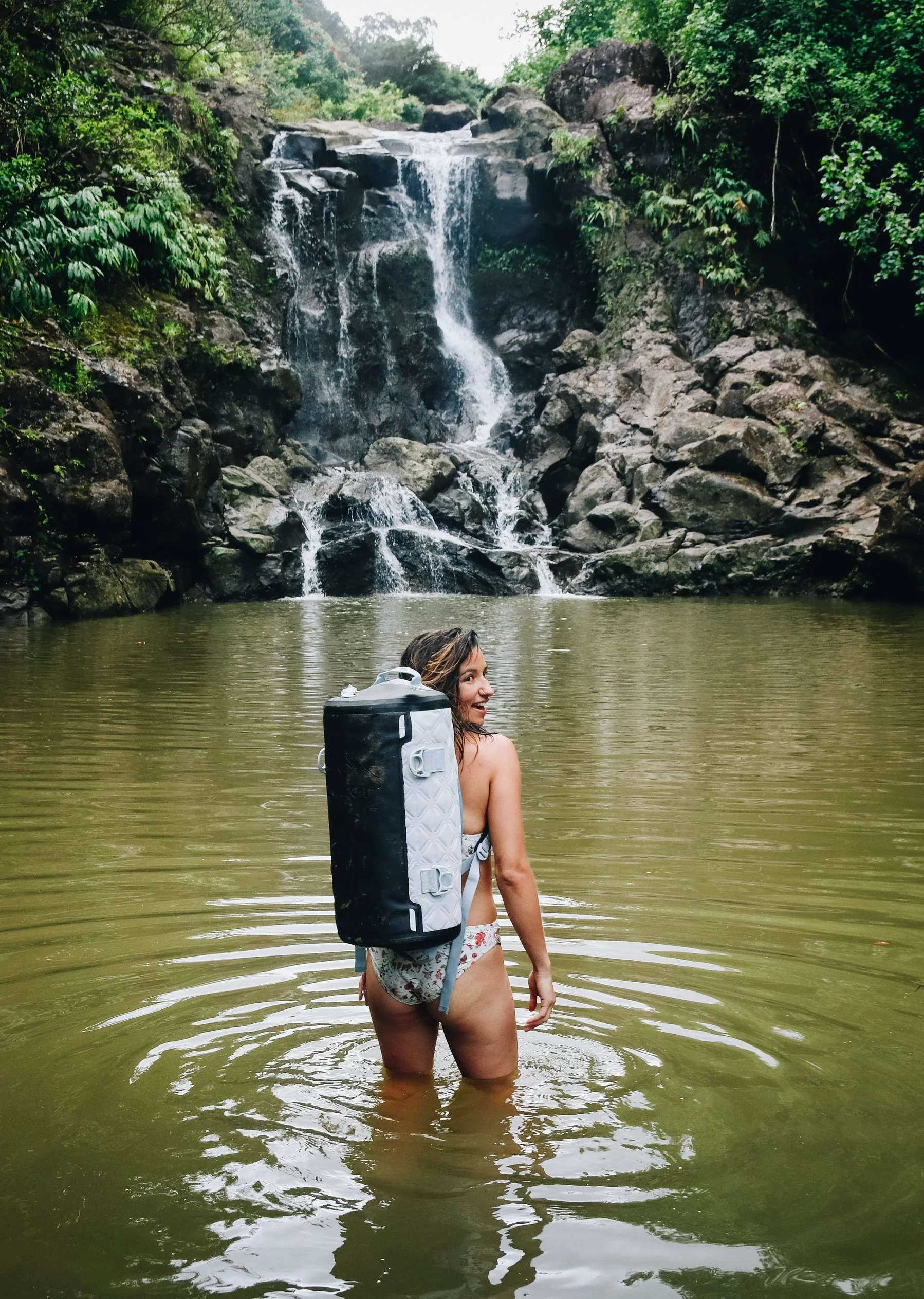 A woman standing waist-deep in a river with a small waterfall in the background, surrounded by lush green trees.