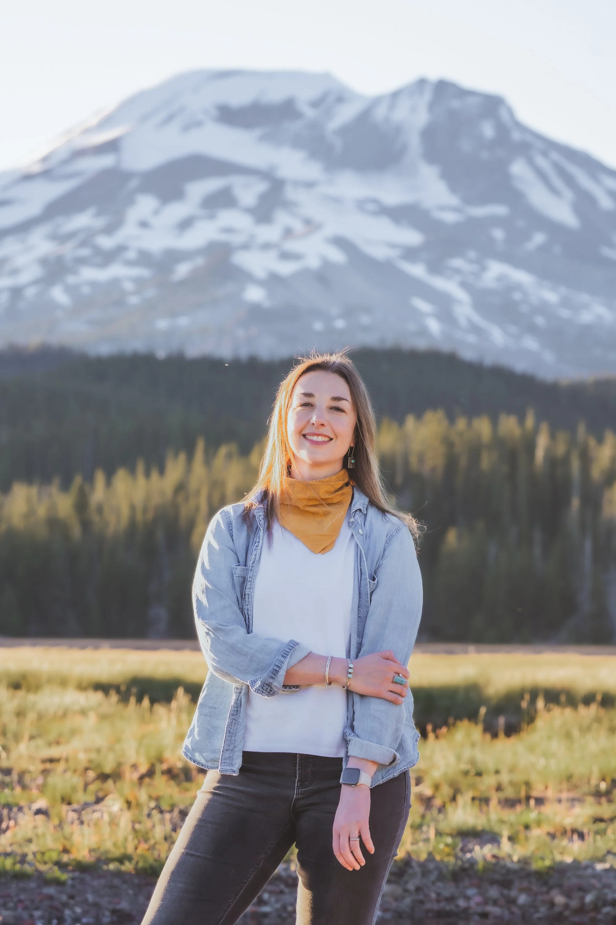 A woman smiles and crosses her arms in front of a mountain landscape with snow-capped peaks and a forested valley.