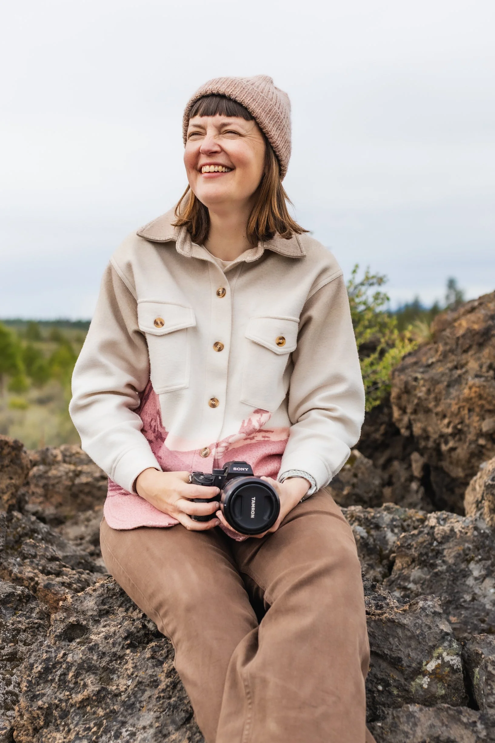 A woman sitting outdoors on rocks, smiling while holding a camera, wearing a beige jacket, brown pants, and a pink knit hat, with a blurry landscape background.