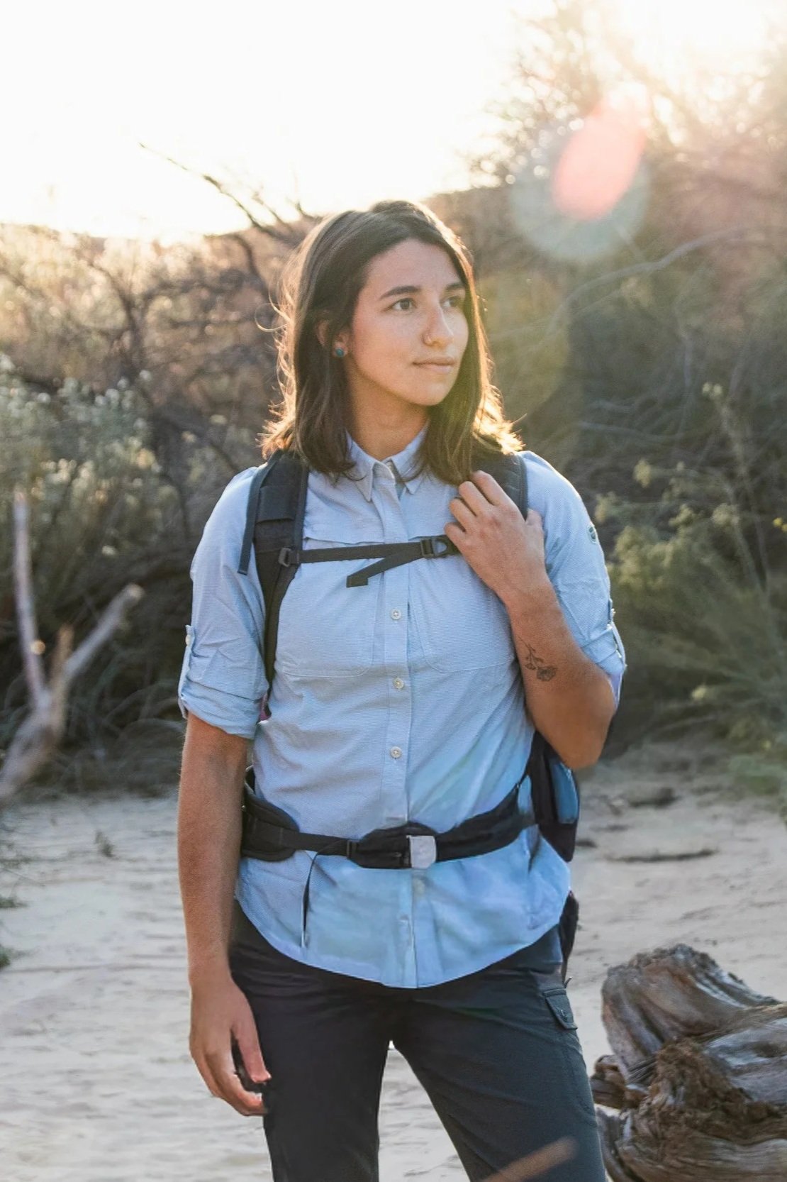 A woman standing outdoors in a natural setting with trees and driftwood, wearing a light blue shirt, black pants, and a backpack, illuminated by sunlight.