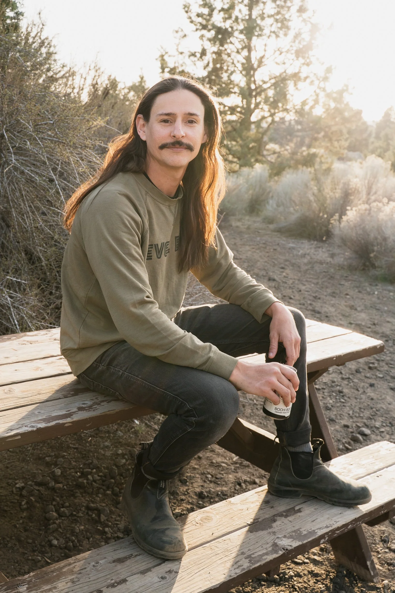 A man with long brown hair, a mustache, wearing a beige sweatshirt and black boots, sitting on a wooden bench outdoors with a drink in his hand, in natural setting with trees and sunlight in the background.