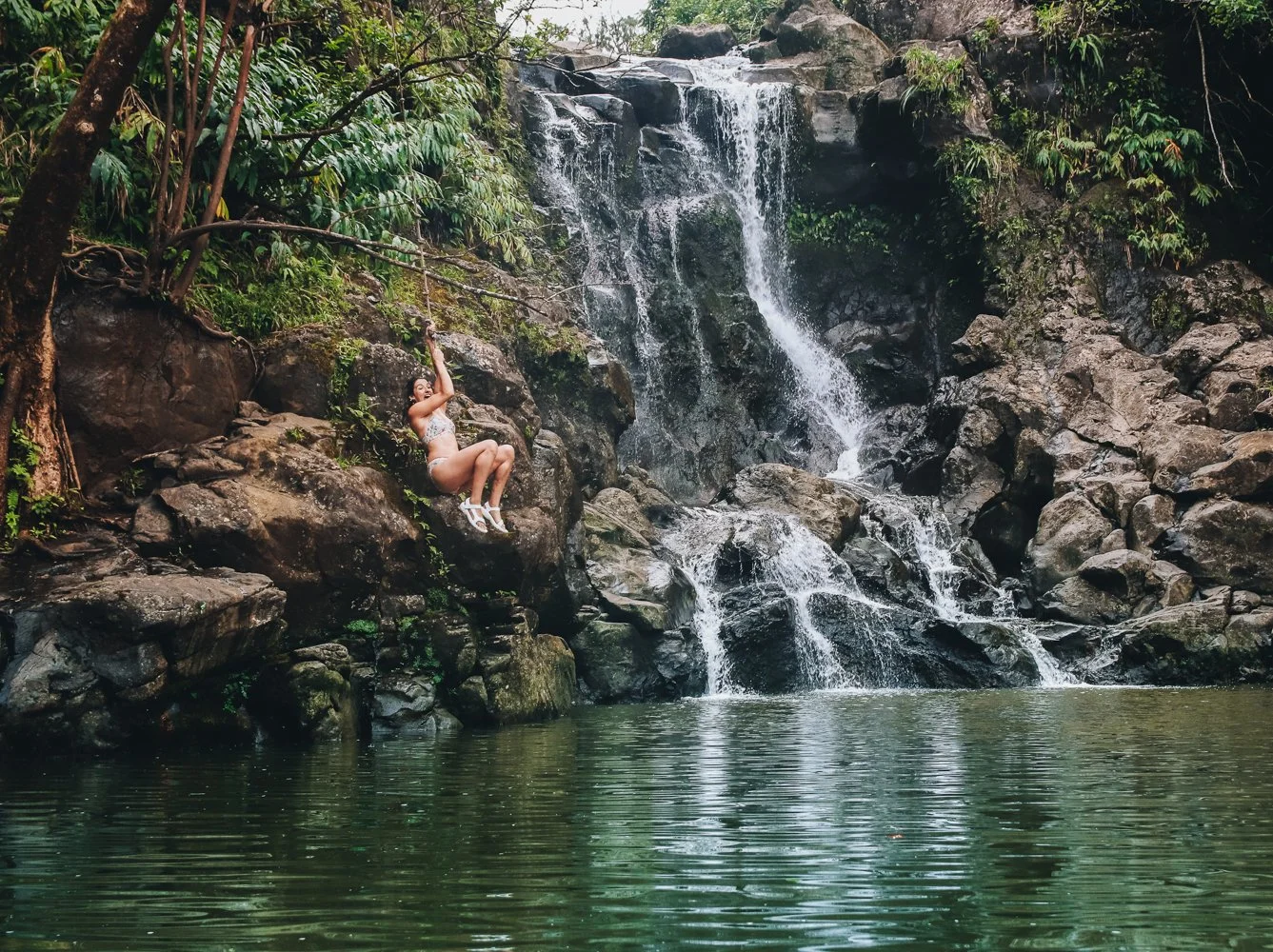 A woman in a swimsuit and white sandals hanging from a green vine beside a waterfall in a jungle setting.