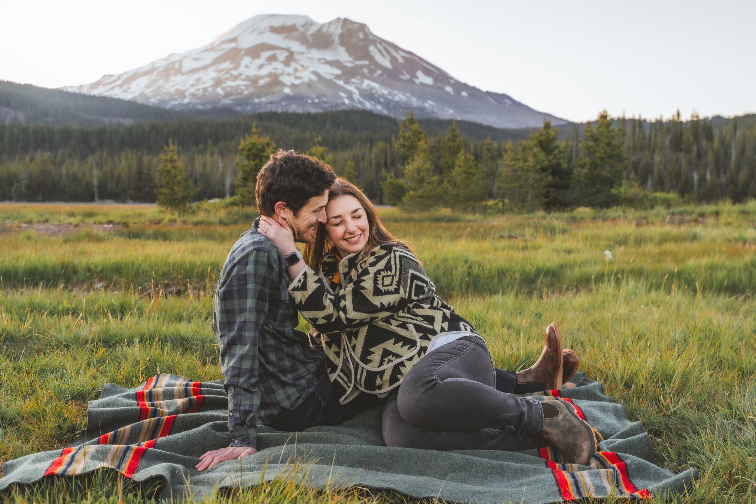 a man and woman snuggle on a blanket in front of a mountain.