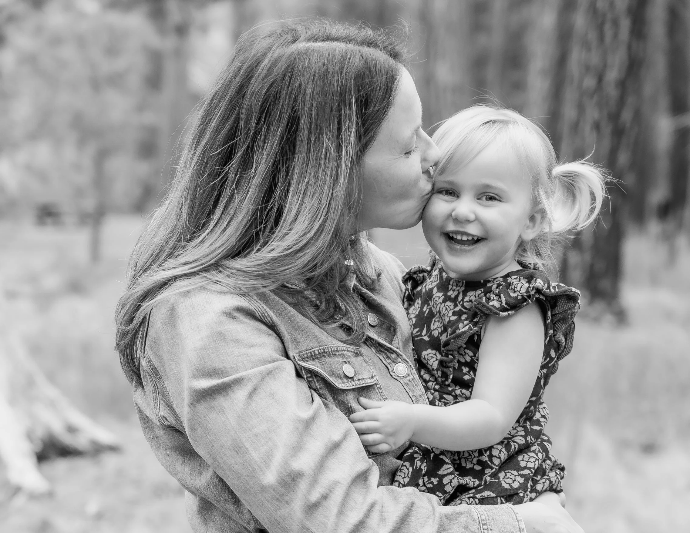 A woman with long hair kissing a young girl with light hair styled in pigtails, both smiling and hugging outdoors with blurred trees in the background.
