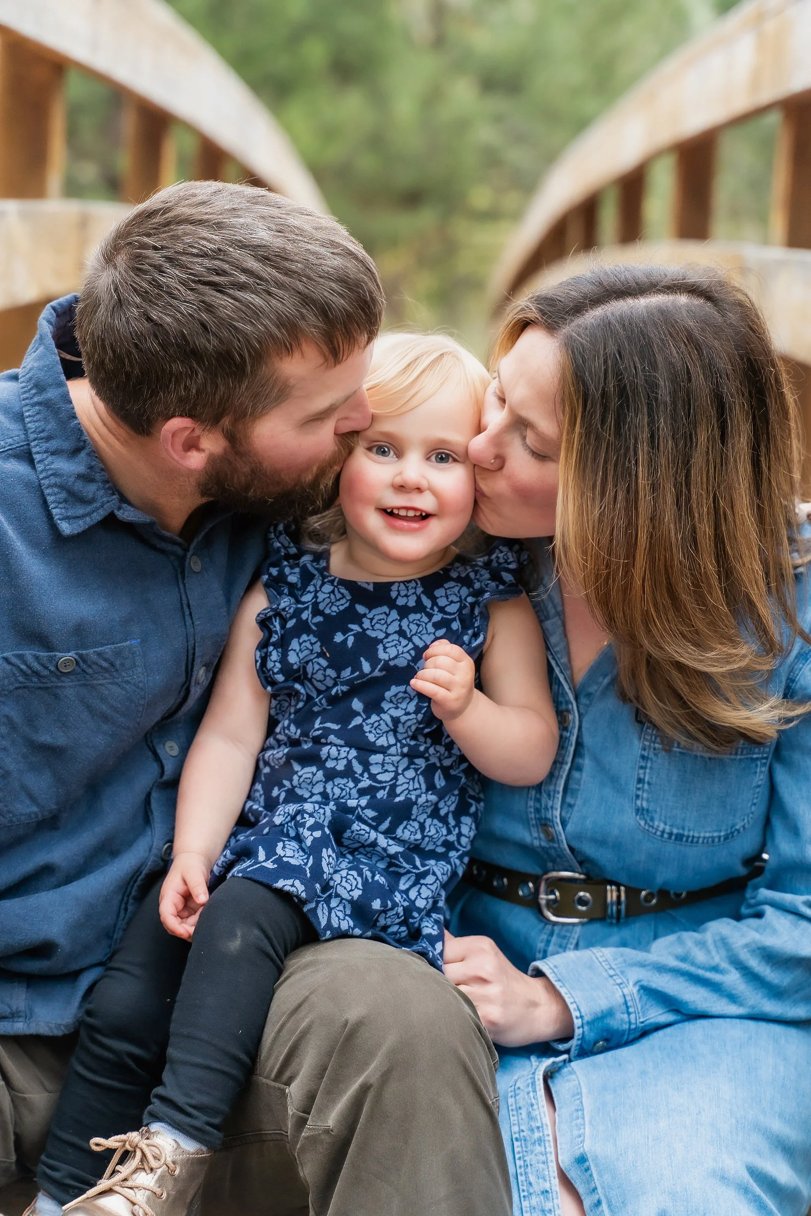 A young girl with blonde hair and blue eyes smiling while being kissed on both cheeks by a man and a woman, likely her parents, on a wooden bridge in a forested area.