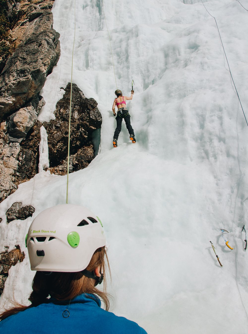 A woman ice climbing on a frozen waterfall, with another person observing at the bottom, both wearing helmets and harnesses, on a sunny outdoor mountain setting.