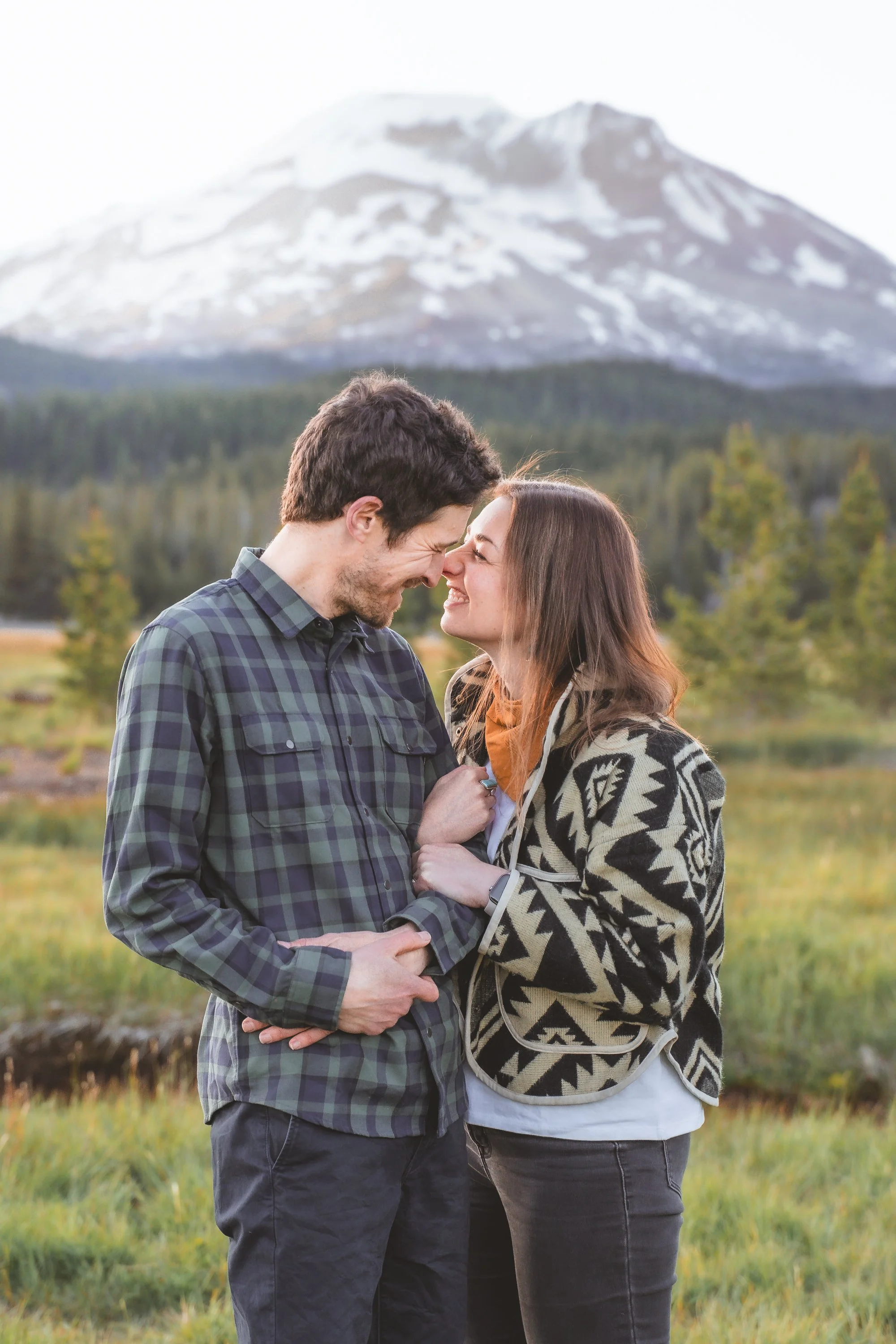 A couple is smiling and touching foreheads outdoors with a mountain in the background. They are dressed casually, and the woman is wearing a patterned jacket.