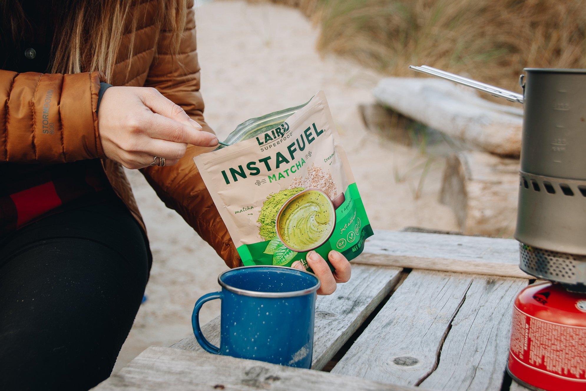 Person opening a bag of LaCroix Matcha flavored instant drink mix while sitting at a rustic outdoor wooden table with camping gear and a blue mug nearby.