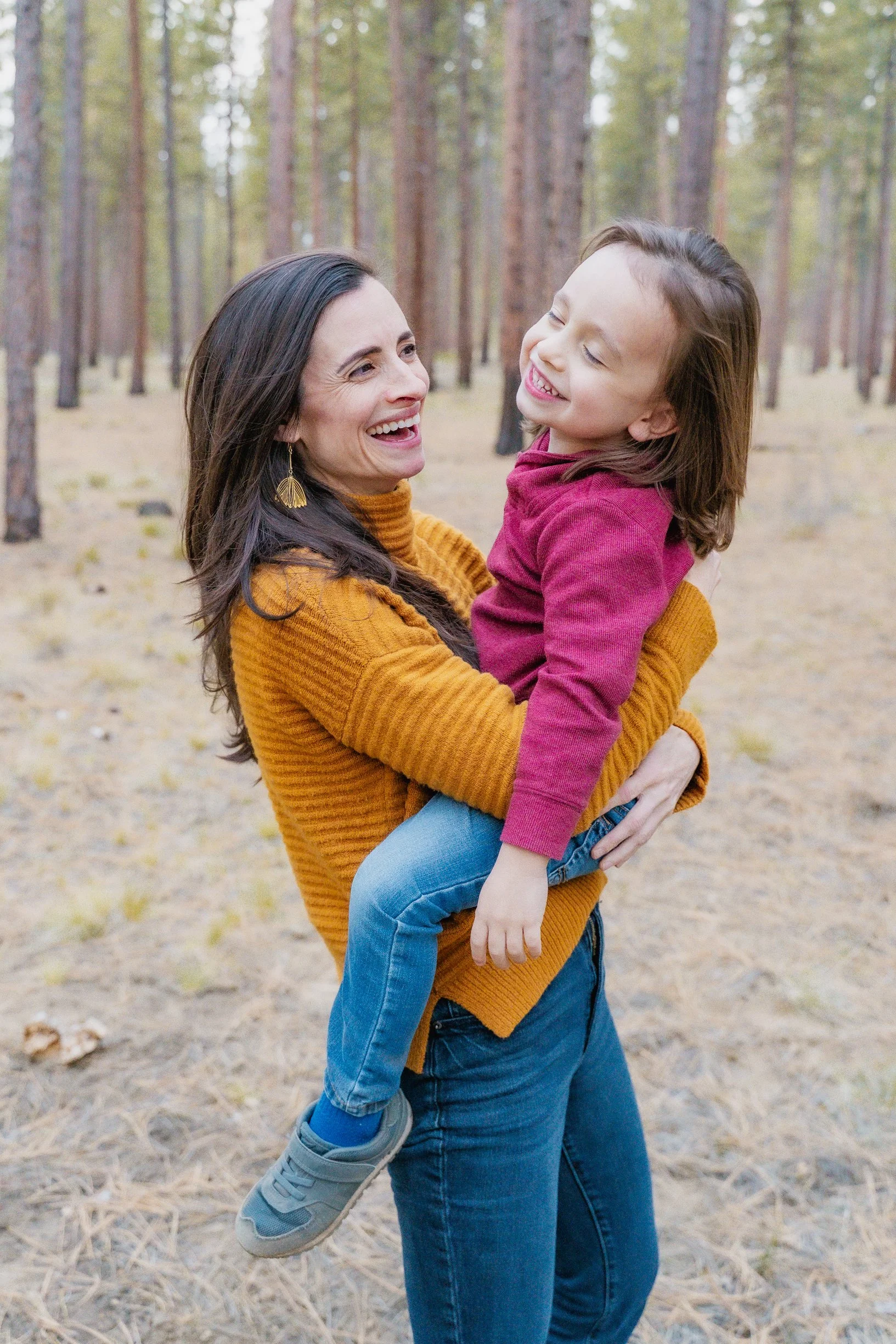 A woman carrying a smiling young girl in a forest with tall pine trees