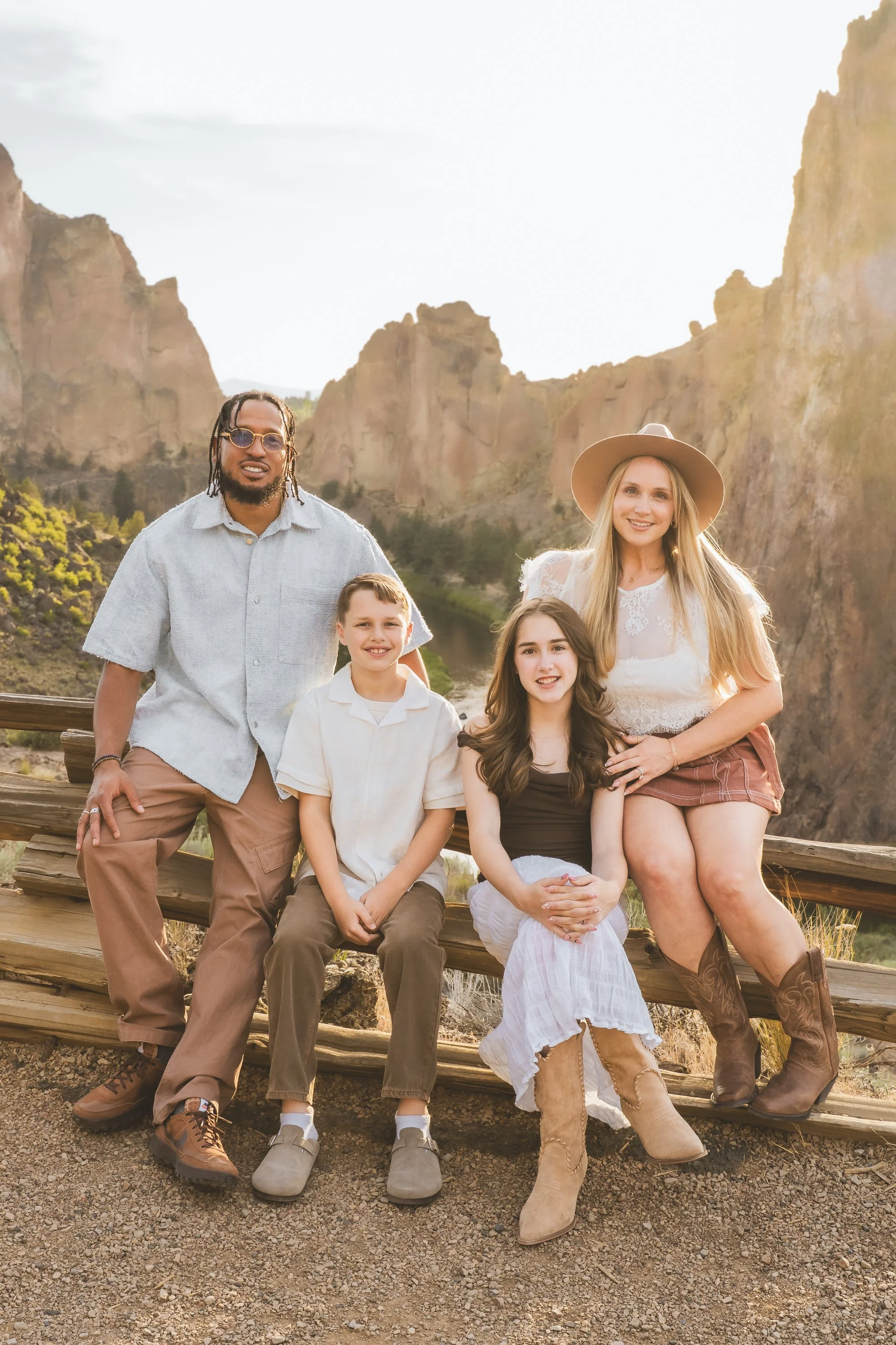 A diverse group of five people, including two adults and three children, standing outdoors in front of a mountain landscape with cliffs and a river, during daylight.