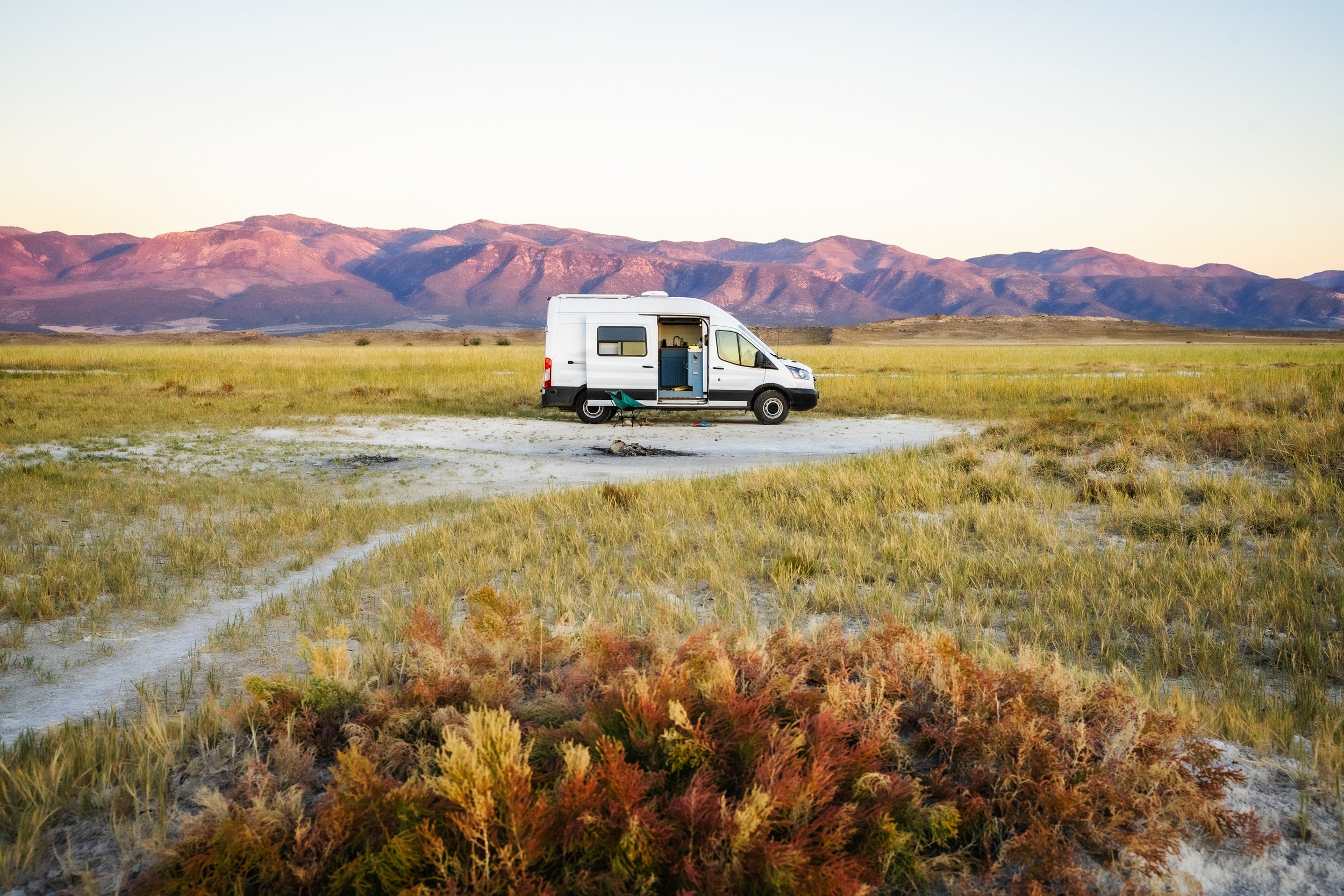 A white camper van parked on a grassy plain with mountains in the background during sunset.