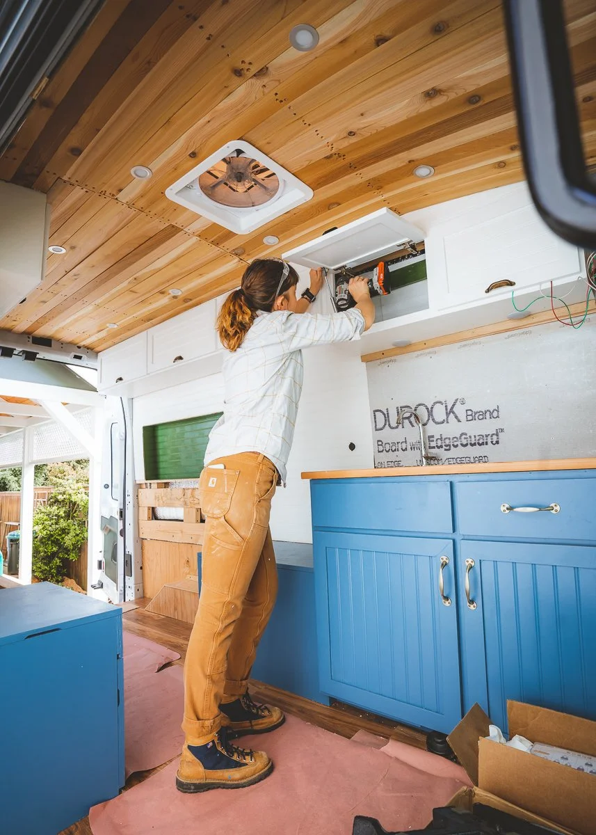 A person installing or repairing a ceiling vent or light fixture inside a wooden-paneled room, using a power drill, with blue cabinetry and a wooden countertop visible.