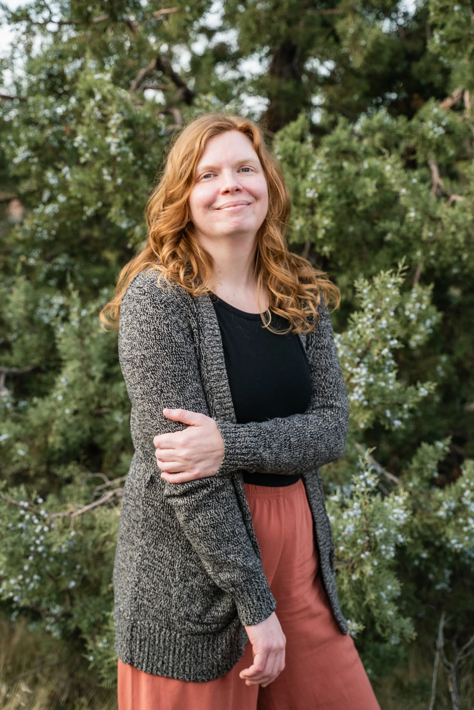 A woman with red hair smiling and standing outdoors with trees in the background. She is wearing a gray knitted cardigan over a black top and rust-colored pants, with one arm crossed over her chest.