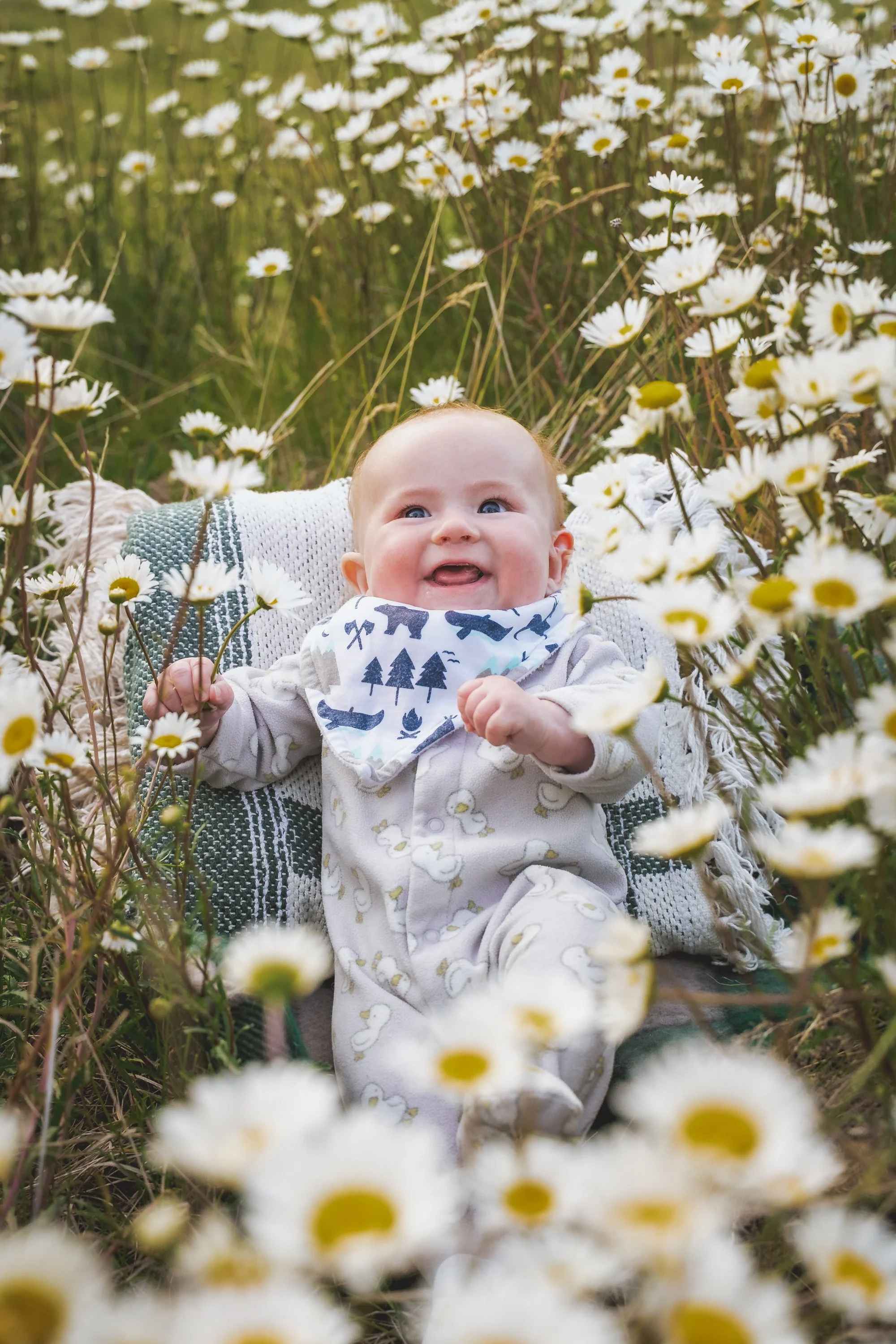 A baby with blue eyes and light hair smiling and lying among daisies in a field.