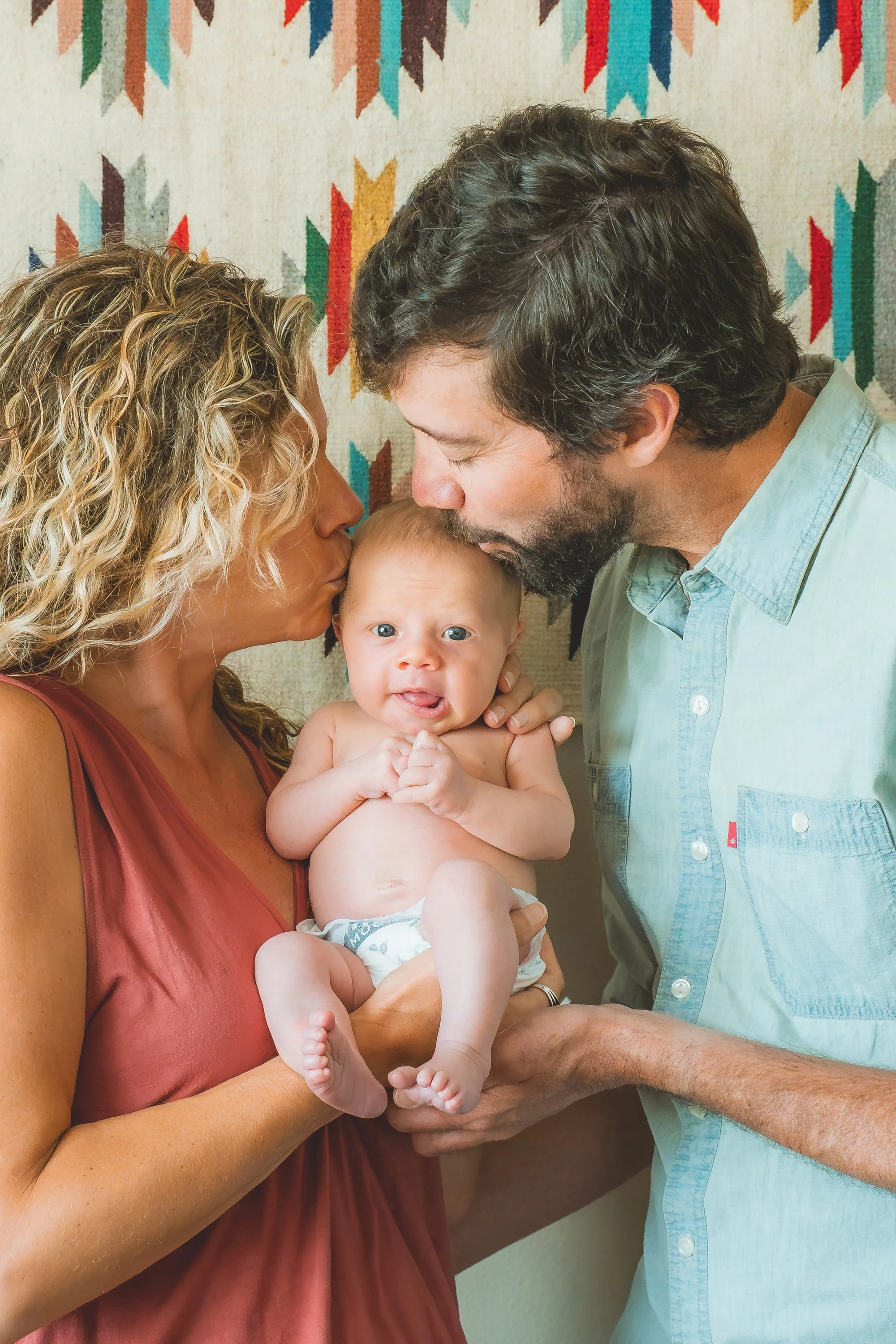A couple with light skin kissing their baby on the forehead against a colorful geometric patterned background.