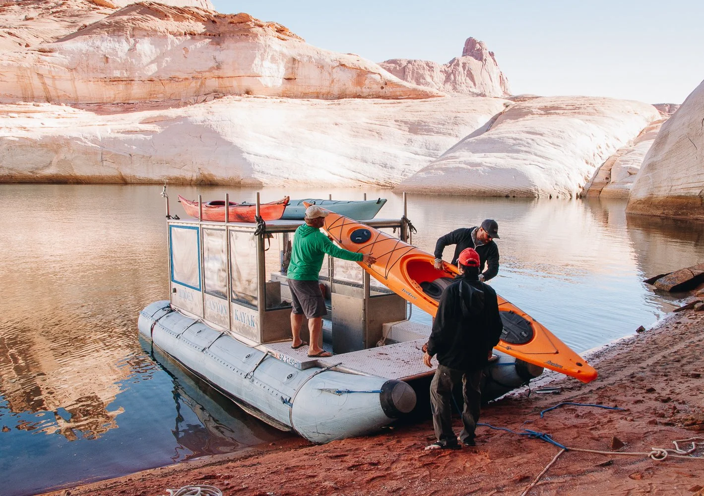 Three men preparing a paddle boat with orange, red, and blue kayaks on a floating platform at the edge of a river, with sandstone cliffs in the background.