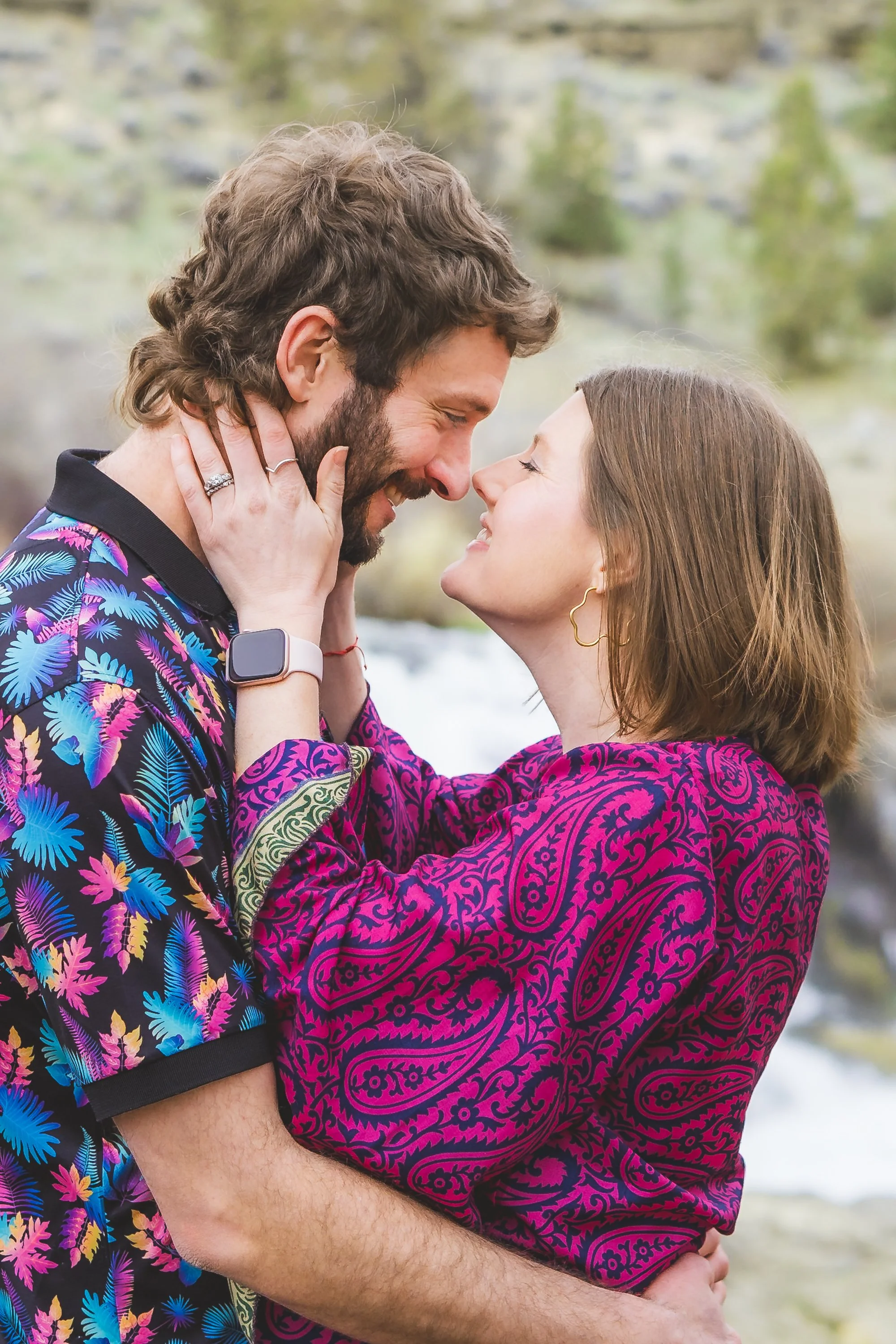 A young couple is smiling and about to kiss outdoors, with a blurred natural landscape in the background. They are dressed in colorful, patterned clothing.