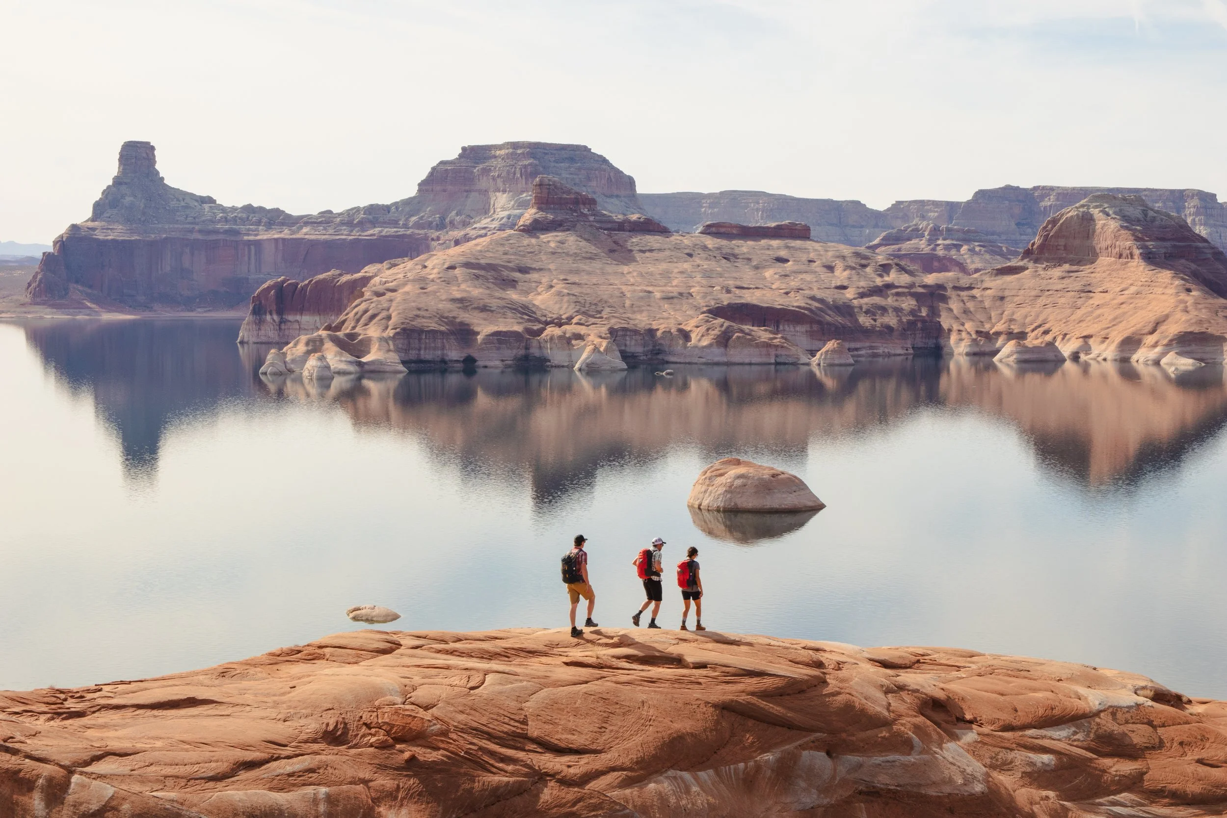 Four hikers with backpacks walk along the rocky shoreline of a calm body of water with large rock formations and mesas in the background at the Grand Canyon.