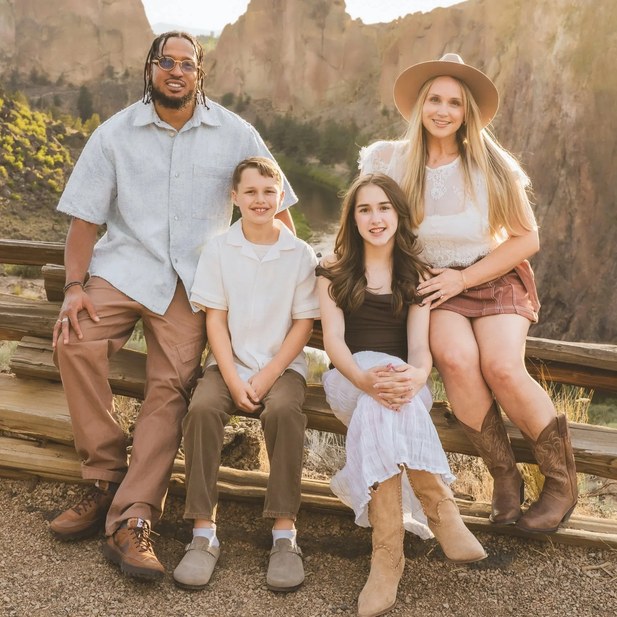 A diverse family group of five people, including two children and three adults, standing outdoors on a wooden bridge with a canyon and a river in the background, during sunset.