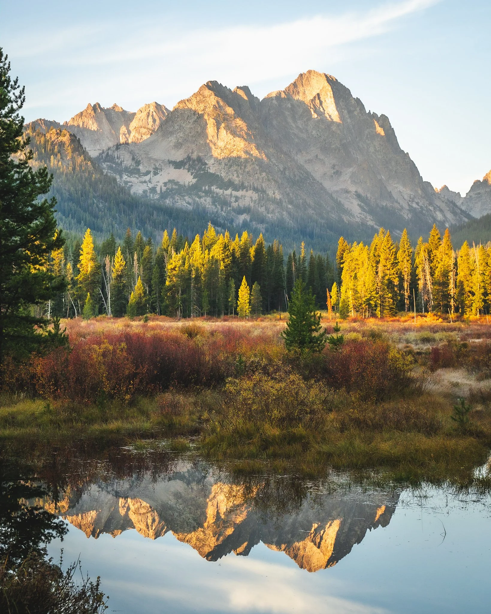 Sunlit mountain range with rugged peaks reflected in a calm lake, surrounded by dense forest and foliage.