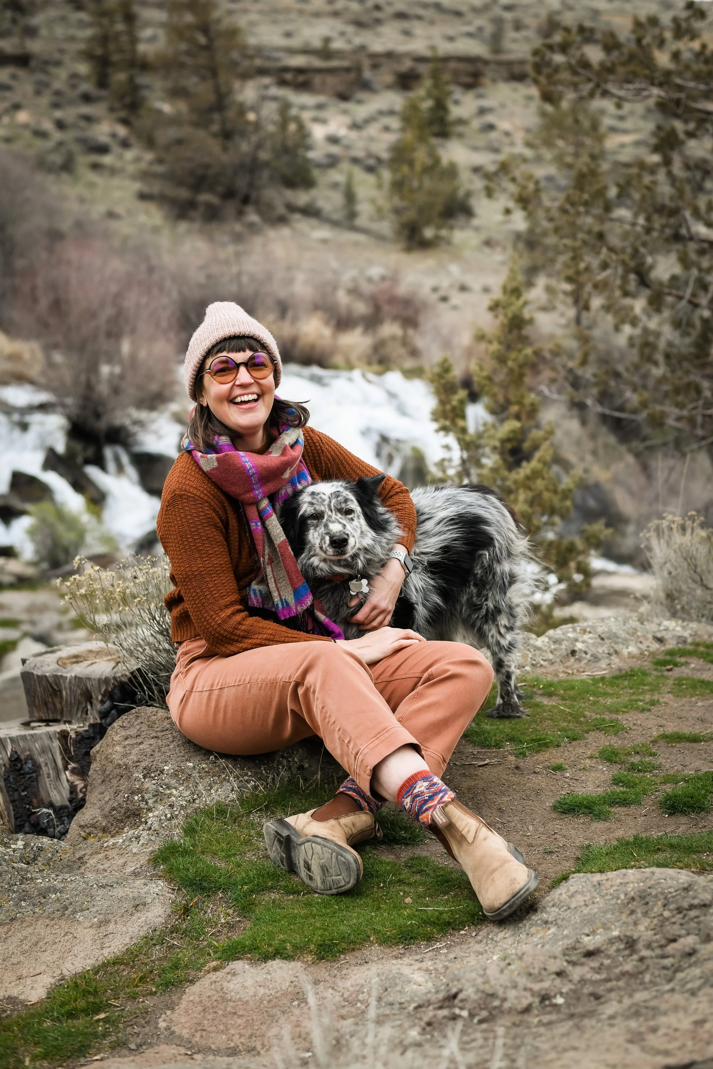 A woman sitting on a log outdoors in front of a river with trees and rocks, smiling and holding a black and white dog.
