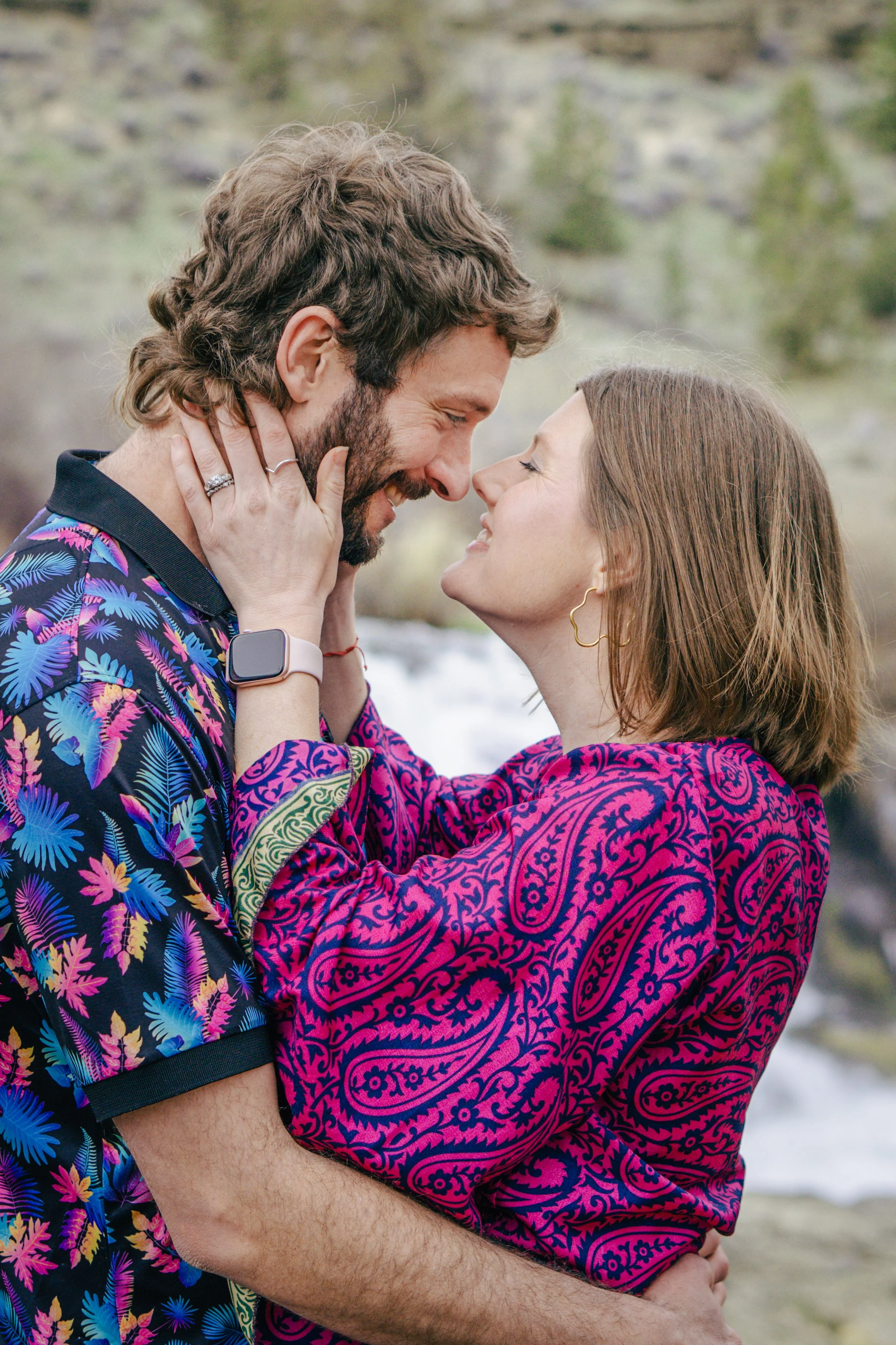 A smiling man and woman are close together, smiling, with their noses almost touching. The woman has her hands on the man's face, and they are outdoors with blurred natural background.