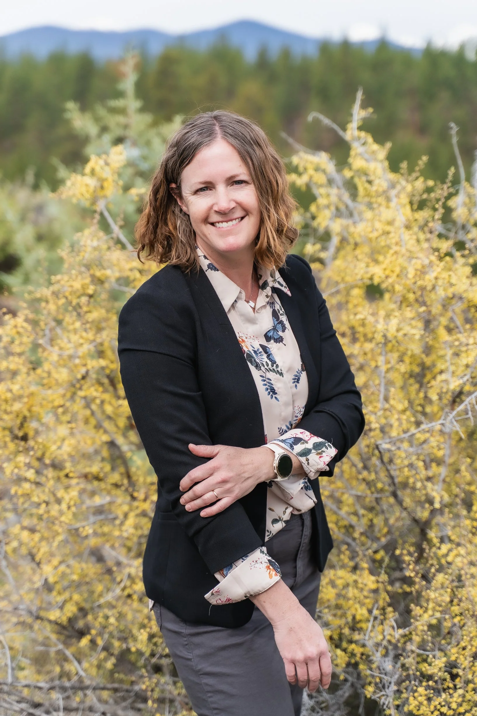A woman with curly brown hair smiling outdoors, standing in front of yellow autumn foliage and distant mountains.