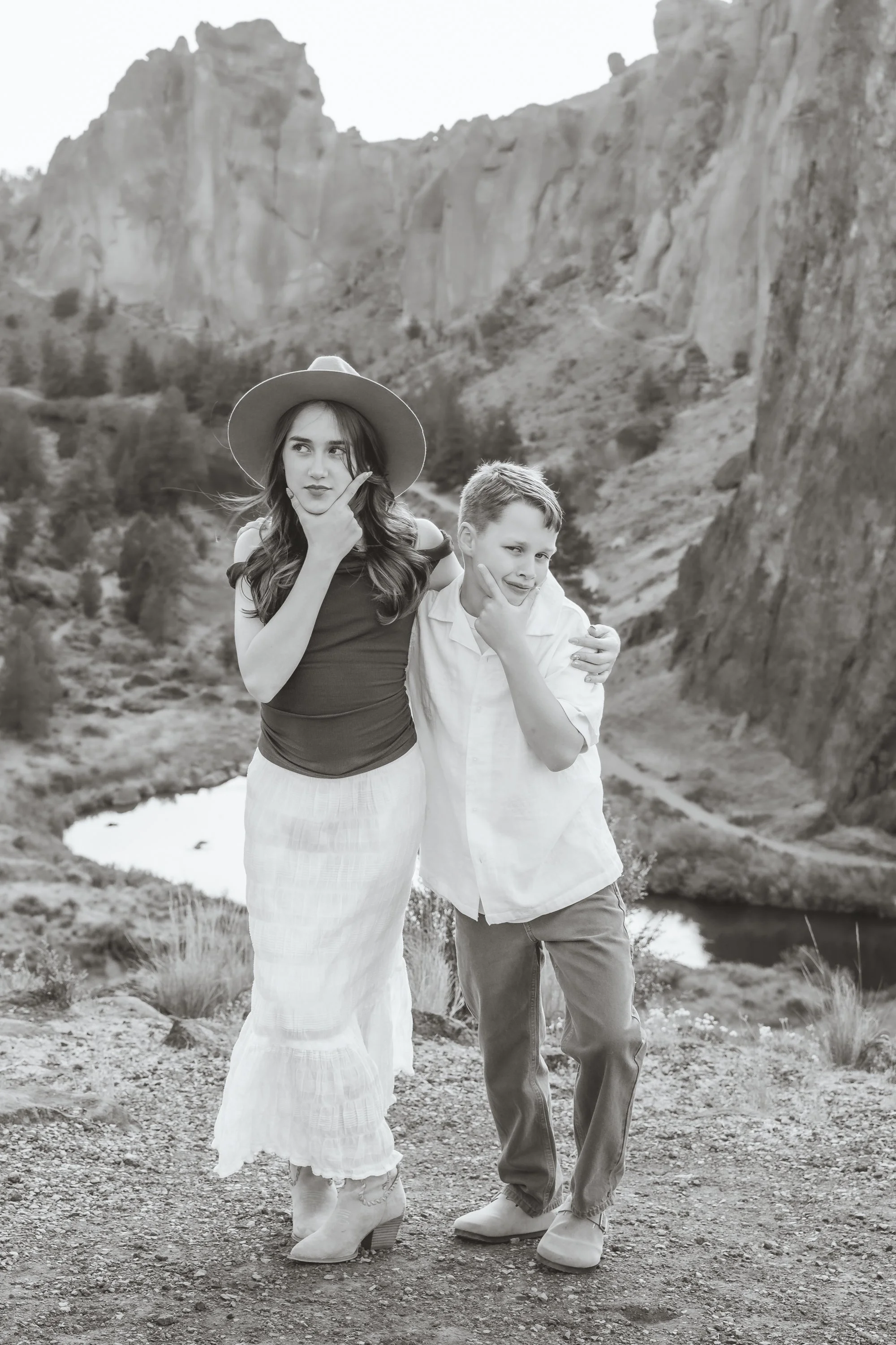 A woman in a wide-brimmed hat and a skirt stands next to a boy in casual clothing, both striking thoughtful poses with their hands on their chins. They are outdoors near a river with steep cliffs in the background, in a rocky, natural landscape.