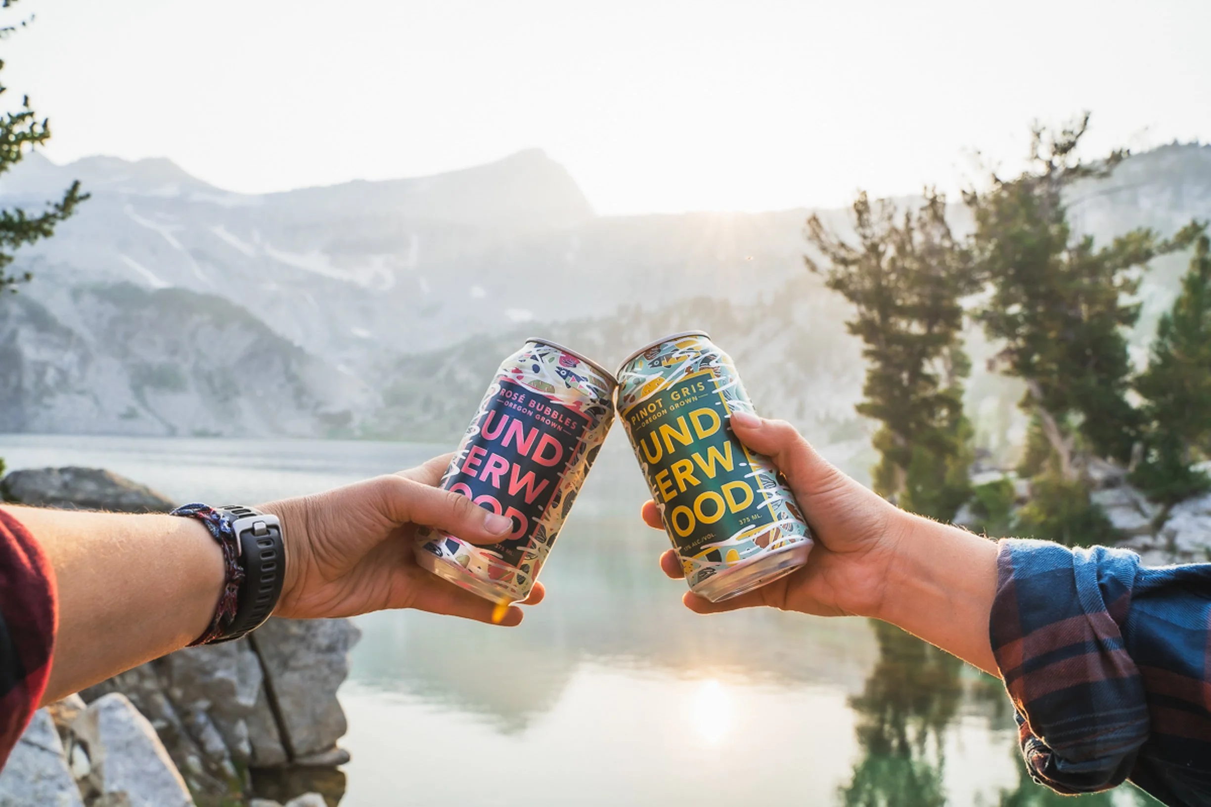 Two people holding cans of wine toasting outdoors by a lake or river in a mountainous area during sunset.