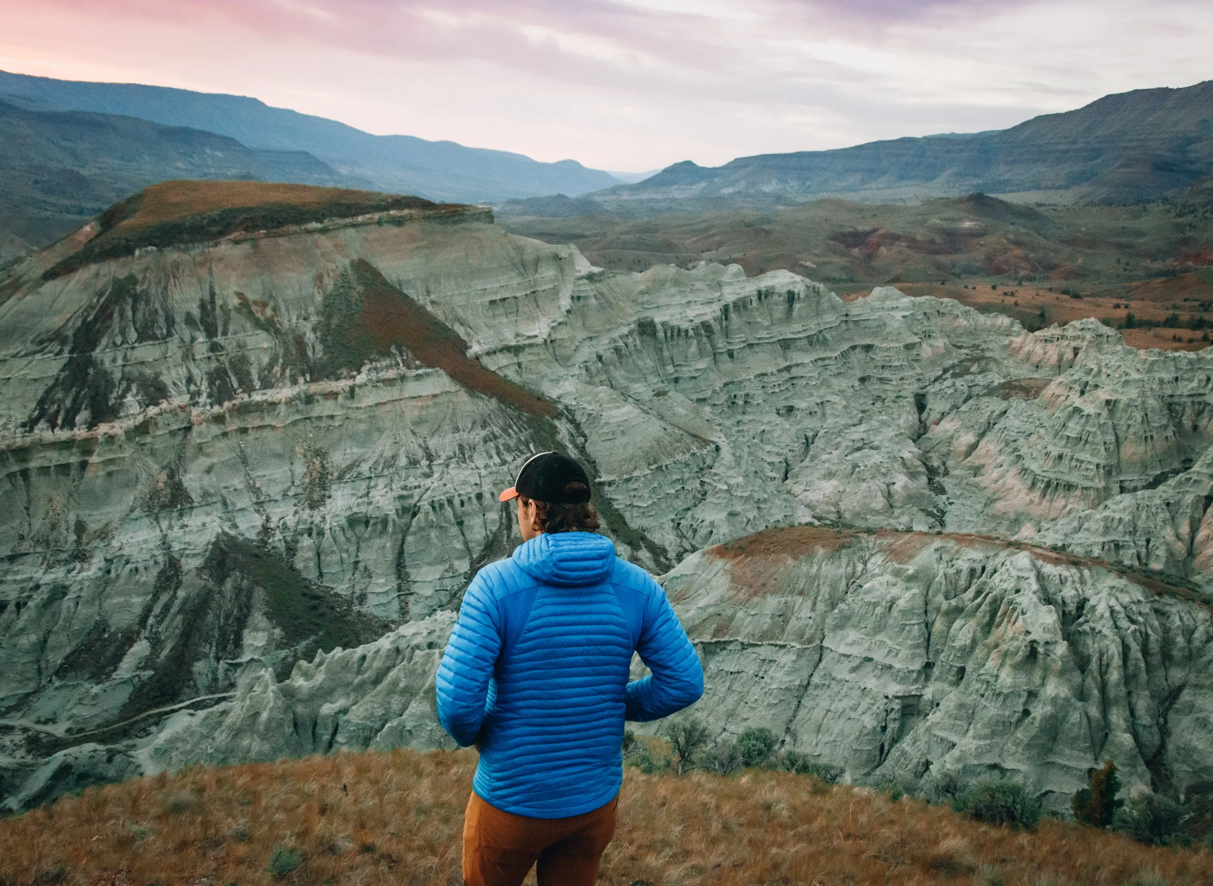 A person in a blue jacket and brown pants standing outdoors in front of a rugged, mountainous landscape with layered rock formations and distant mountains under a cloudy sky.